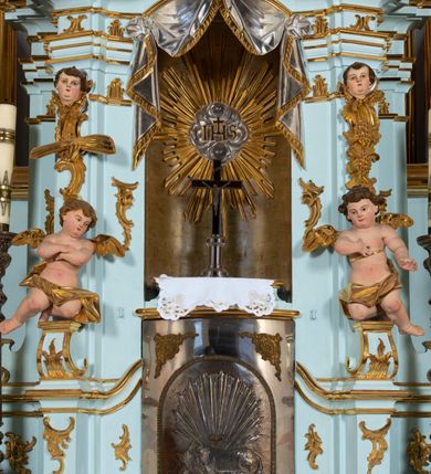 Zdjęcie nr 21: Nastawa architektoniczna, jednoosiowa, jednokondygnacyjna, ustawiona na wysokim cokole. 
Ołtarz drewniany, prostopadłościenny. Na nim tabernakulum z tronem wystawienia. Tabernakulum w formie drewnianej, prostopadłościennej skrzynki, obitej blachą, o wypukłej ściance frontowej, zamkniętej profilowanym gzymsem. Drzwiczki w formie stojącego prostokąta zamkniętego łukiem półkolistym, obwiedzione ramą, z wizerunkiem baranka z krzyżem, leżącego na księdze z pieczęciami, w glorii. Tabernakulum w drewnianej obudowie, wpisane w wysoki cokół, na nim tron wystawienia w formie obrotowej niszy w kształcie stojącego prostokąta, z podwieszoną draperią. Nisza flankowana parą pilastrów, przechodzących w spływy wolutowe, na których siedzą putta. Pilastry dźwigają pełne belkowanie, wyłamane na ich osi, częściowo przerwane przez niszę. Pilastry dekorowane uskrzydlonymi główkami anielskimi oraz snopkiem zboża i kiściami winogron. Cały tron wystawienia dekorowany rocaille&#039;m. 
Nastawa ujęta dwiema parami kolumn o kapitelach kompozytowych, dźwigających pełne belkowanie, wyłamane pomiędzy nimi, przerwane nad polem środkowym. Na belkowaniu fragmenty przerwanego, półkolistego przyczółka, na których leżą putta. Całe pole środkowe wypełnia wnęka w formie stojącego prostokąta zamkniętego łukiem półkolistym, ujęta profilowaną ramą, z figurą Chrystusa Ukrzyżowanego. Nad łukiem wnęki, pomiędzy obłokami, gołębica Ducha Świętego w glorii promienistej. Pomiędzy kolumnami płyciny z uskrzydlonymi główkami anielskimi na obłokach, rocaille oraz figury św.  Jana Ewangelisty i Matki Boskiej na rocaille&#039;owych wspornikach. Kondygnacja ujęta uszami utworzonymi z rocaille&#039;u. Nastawa zwieńczona rzeźbą tronującego Boga Ojca z globem i berłem, pośród obłoków i uskrzydlonych główek anielskich, w glorii promienistej. 
Struktura polichromowana w kolorze błękitnym, kolumny, profile i ornamentyka złocone. 
