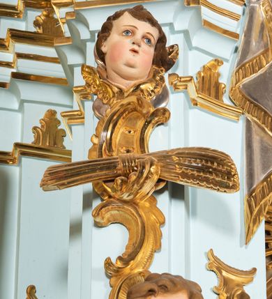 Zdjęcie nr 31: Nastawa architektoniczna, jednoosiowa, jednokondygnacyjna, ustawiona na wysokim cokole. 
Ołtarz drewniany, prostopadłościenny. Na nim tabernakulum z tronem wystawienia. Tabernakulum w formie drewnianej, prostopadłościennej skrzynki, obitej blachą, o wypukłej ściance frontowej, zamkniętej profilowanym gzymsem. Drzwiczki w formie stojącego prostokąta zamkniętego łukiem półkolistym, obwiedzione ramą, z wizerunkiem baranka z krzyżem, leżącego na księdze z pieczęciami, w glorii. Tabernakulum w drewnianej obudowie, wpisane w wysoki cokół, na nim tron wystawienia w formie obrotowej niszy w kształcie stojącego prostokąta, z podwieszoną draperią. Nisza flankowana parą pilastrów, przechodzących w spływy wolutowe, na których siedzą putta. Pilastry dźwigają pełne belkowanie, wyłamane na ich osi, częściowo przerwane przez niszę. Pilastry dekorowane uskrzydlonymi główkami anielskimi oraz snopkiem zboża i kiściami winogron. Cały tron wystawienia dekorowany rocaille&#039;m. 
Nastawa ujęta dwiema parami kolumn o kapitelach kompozytowych, dźwigających pełne belkowanie, wyłamane pomiędzy nimi, przerwane nad polem środkowym. Na belkowaniu fragmenty przerwanego, półkolistego przyczółka, na których leżą putta. Całe pole środkowe wypełnia wnęka w formie stojącego prostokąta zamkniętego łukiem półkolistym, ujęta profilowaną ramą, z figurą Chrystusa Ukrzyżowanego. Nad łukiem wnęki, pomiędzy obłokami, gołębica Ducha Świętego w glorii promienistej. Pomiędzy kolumnami płyciny z uskrzydlonymi główkami anielskimi na obłokach, rocaille oraz figury św.  Jana Ewangelisty i Matki Boskiej na rocaille&#039;owych wspornikach. Kondygnacja ujęta uszami utworzonymi z rocaille&#039;u. Nastawa zwieńczona rzeźbą tronującego Boga Ojca z globem i berłem, pośród obłoków i uskrzydlonych główek anielskich, w glorii promienistej. 
Struktura polichromowana w kolorze błękitnym, kolumny, profile i ornamentyka złocone. 
