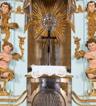 Zdjęcie nr 25: Nastawa architektoniczna, jednoosiowa, jednokondygnacyjna, ustawiona na wysokim cokole. 
Ołtarz drewniany, prostopadłościenny. Na nim tabernakulum z tronem wystawienia. Tabernakulum w formie drewnianej, prostopadłościennej skrzynki, obitej blachą, o wypukłej ściance frontowej, zamkniętej profilowanym gzymsem. Drzwiczki w formie stojącego prostokąta zamkniętego łukiem półkolistym, obwiedzione ramą, z wizerunkiem baranka z krzyżem, leżącego na księdze z pieczęciami, w glorii. Tabernakulum w drewnianej obudowie, wpisane w wysoki cokół, na nim tron wystawienia w formie obrotowej niszy w kształcie stojącego prostokąta, z podwieszoną draperią. Nisza flankowana parą pilastrów, przechodzących w spływy wolutowe, na których siedzą putta. Pilastry dźwigają pełne belkowanie, wyłamane na ich osi, częściowo przerwane przez niszę. Pilastry dekorowane uskrzydlonymi główkami anielskimi oraz snopkiem zboża i kiściami winogron. Cały tron wystawienia dekorowany rocaille&#039;m. 
Nastawa ujęta dwiema parami kolumn o kapitelach kompozytowych, dźwigających pełne belkowanie, wyłamane pomiędzy nimi, przerwane nad polem środkowym. Na belkowaniu fragmenty przerwanego, półkolistego przyczółka, na których leżą putta. Całe pole środkowe wypełnia wnęka w formie stojącego prostokąta zamkniętego łukiem półkolistym, ujęta profilowaną ramą, z figurą Chrystusa Ukrzyżowanego. Nad łukiem wnęki, pomiędzy obłokami, gołębica Ducha Świętego w glorii promienistej. Pomiędzy kolumnami płyciny z uskrzydlonymi główkami anielskimi na obłokach, rocaille oraz figury św.  Jana Ewangelisty i Matki Boskiej na rocaille&#039;owych wspornikach. Kondygnacja ujęta uszami utworzonymi z rocaille&#039;u. Nastawa zwieńczona rzeźbą tronującego Boga Ojca z globem i berłem, pośród obłoków i uskrzydlonych główek anielskich, w glorii promienistej. 
Struktura polichromowana w kolorze błękitnym, kolumny, profile i ornamentyka złocone. 
