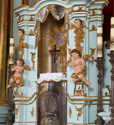 Zdjęcie nr 23: Nastawa architektoniczna, jednoosiowa, jednokondygnacyjna, ustawiona na wysokim cokole. 
Ołtarz drewniany, prostopadłościenny. Na nim tabernakulum z tronem wystawienia. Tabernakulum w formie drewnianej, prostopadłościennej skrzynki, obitej blachą, o wypukłej ściance frontowej, zamkniętej profilowanym gzymsem. Drzwiczki w formie stojącego prostokąta zamkniętego łukiem półkolistym, obwiedzione ramą, z wizerunkiem baranka z krzyżem, leżącego na księdze z pieczęciami, w glorii. Tabernakulum w drewnianej obudowie, wpisane w wysoki cokół, na nim tron wystawienia w formie obrotowej niszy w kształcie stojącego prostokąta, z podwieszoną draperią. Nisza flankowana parą pilastrów, przechodzących w spływy wolutowe, na których siedzą putta. Pilastry dźwigają pełne belkowanie, wyłamane na ich osi, częściowo przerwane przez niszę. Pilastry dekorowane uskrzydlonymi główkami anielskimi oraz snopkiem zboża i kiściami winogron. Cały tron wystawienia dekorowany rocaille&#039;m. 
Nastawa ujęta dwiema parami kolumn o kapitelach kompozytowych, dźwigających pełne belkowanie, wyłamane pomiędzy nimi, przerwane nad polem środkowym. Na belkowaniu fragmenty przerwanego, półkolistego przyczółka, na których leżą putta. Całe pole środkowe wypełnia wnęka w formie stojącego prostokąta zamkniętego łukiem półkolistym, ujęta profilowaną ramą, z figurą Chrystusa Ukrzyżowanego. Nad łukiem wnęki, pomiędzy obłokami, gołębica Ducha Świętego w glorii promienistej. Pomiędzy kolumnami płyciny z uskrzydlonymi główkami anielskimi na obłokach, rocaille oraz figury św.  Jana Ewangelisty i Matki Boskiej na rocaille&#039;owych wspornikach. Kondygnacja ujęta uszami utworzonymi z rocaille&#039;u. Nastawa zwieńczona rzeźbą tronującego Boga Ojca z globem i berłem, pośród obłoków i uskrzydlonych główek anielskich, w glorii promienistej. 
Struktura polichromowana w kolorze błękitnym, kolumny, profile i ornamentyka złocone. 
