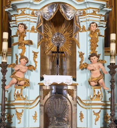 Zdjęcie nr 20: Nastawa architektoniczna, jednoosiowa, jednokondygnacyjna, ustawiona na wysokim cokole. 
Ołtarz drewniany, prostopadłościenny. Na nim tabernakulum z tronem wystawienia. Tabernakulum w formie drewnianej, prostopadłościennej skrzynki, obitej blachą, o wypukłej ściance frontowej, zamkniętej profilowanym gzymsem. Drzwiczki w formie stojącego prostokąta zamkniętego łukiem półkolistym, obwiedzione ramą, z wizerunkiem baranka z krzyżem, leżącego na księdze z pieczęciami, w glorii. Tabernakulum w drewnianej obudowie, wpisane w wysoki cokół, na nim tron wystawienia w formie obrotowej niszy w kształcie stojącego prostokąta, z podwieszoną draperią. Nisza flankowana parą pilastrów, przechodzących w spływy wolutowe, na których siedzą putta. Pilastry dźwigają pełne belkowanie, wyłamane na ich osi, częściowo przerwane przez niszę. Pilastry dekorowane uskrzydlonymi główkami anielskimi oraz snopkiem zboża i kiściami winogron. Cały tron wystawienia dekorowany rocaille&#039;m. 
Nastawa ujęta dwiema parami kolumn o kapitelach kompozytowych, dźwigających pełne belkowanie, wyłamane pomiędzy nimi, przerwane nad polem środkowym. Na belkowaniu fragmenty przerwanego, półkolistego przyczółka, na których leżą putta. Całe pole środkowe wypełnia wnęka w formie stojącego prostokąta zamkniętego łukiem półkolistym, ujęta profilowaną ramą, z figurą Chrystusa Ukrzyżowanego. Nad łukiem wnęki, pomiędzy obłokami, gołębica Ducha Świętego w glorii promienistej. Pomiędzy kolumnami płyciny z uskrzydlonymi główkami anielskimi na obłokach, rocaille oraz figury św.  Jana Ewangelisty i Matki Boskiej na rocaille&#039;owych wspornikach. Kondygnacja ujęta uszami utworzonymi z rocaille&#039;u. Nastawa zwieńczona rzeźbą tronującego Boga Ojca z globem i berłem, pośród obłoków i uskrzydlonych główek anielskich, w glorii promienistej. 
Struktura polichromowana w kolorze błękitnym, kolumny, profile i ornamentyka złocone. 
