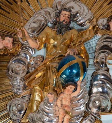 Zdjęcie nr 52: Nastawa architektoniczna, jednoosiowa, jednokondygnacyjna, ustawiona na wysokim cokole. 
Ołtarz drewniany, prostopadłościenny. Na nim tabernakulum z tronem wystawienia. Tabernakulum w formie drewnianej, prostopadłościennej skrzynki, obitej blachą, o wypukłej ściance frontowej, zamkniętej profilowanym gzymsem. Drzwiczki w formie stojącego prostokąta zamkniętego łukiem półkolistym, obwiedzione ramą, z wizerunkiem baranka z krzyżem, leżącego na księdze z pieczęciami, w glorii. Tabernakulum w drewnianej obudowie, wpisane w wysoki cokół, na nim tron wystawienia w formie obrotowej niszy w kształcie stojącego prostokąta, z podwieszoną draperią. Nisza flankowana parą pilastrów, przechodzących w spływy wolutowe, na których siedzą putta. Pilastry dźwigają pełne belkowanie, wyłamane na ich osi, częściowo przerwane przez niszę. Pilastry dekorowane uskrzydlonymi główkami anielskimi oraz snopkiem zboża i kiściami winogron. Cały tron wystawienia dekorowany rocaille&#039;m. 
Nastawa ujęta dwiema parami kolumn o kapitelach kompozytowych, dźwigających pełne belkowanie, wyłamane pomiędzy nimi, przerwane nad polem środkowym. Na belkowaniu fragmenty przerwanego, półkolistego przyczółka, na których leżą putta. Całe pole środkowe wypełnia wnęka w formie stojącego prostokąta zamkniętego łukiem półkolistym, ujęta profilowaną ramą, z figurą Chrystusa Ukrzyżowanego. Nad łukiem wnęki, pomiędzy obłokami, gołębica Ducha Świętego w glorii promienistej. Pomiędzy kolumnami płyciny z uskrzydlonymi główkami anielskimi na obłokach, rocaille oraz figury św.  Jana Ewangelisty i Matki Boskiej na rocaille&#039;owych wspornikach. Kondygnacja ujęta uszami utworzonymi z rocaille&#039;u. Nastawa zwieńczona rzeźbą tronującego Boga Ojca z globem i berłem, pośród obłoków i uskrzydlonych główek anielskich, w glorii promienistej. 
Struktura polichromowana w kolorze błękitnym, kolumny, profile i ornamentyka złocone. 
