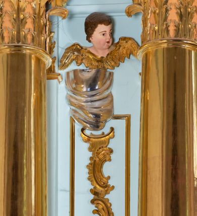 Zdjęcie nr 29: Nastawa architektoniczna, jednoosiowa, jednokondygnacyjna, ustawiona na wysokim cokole. 
Ołtarz drewniany, prostopadłościenny. Na nim tabernakulum z tronem wystawienia. Tabernakulum w formie drewnianej, prostopadłościennej skrzynki, obitej blachą, o wypukłej ściance frontowej, zamkniętej profilowanym gzymsem. Drzwiczki w formie stojącego prostokąta zamkniętego łukiem półkolistym, obwiedzione ramą, z wizerunkiem baranka z krzyżem, leżącego na księdze z pieczęciami, w glorii. Tabernakulum w drewnianej obudowie, wpisane w wysoki cokół, na nim tron wystawienia w formie obrotowej niszy w kształcie stojącego prostokąta, z podwieszoną draperią. Nisza flankowana parą pilastrów, przechodzących w spływy wolutowe, na których siedzą putta. Pilastry dźwigają pełne belkowanie, wyłamane na ich osi, częściowo przerwane przez niszę. Pilastry dekorowane uskrzydlonymi główkami anielskimi oraz snopkiem zboża i kiściami winogron. Cały tron wystawienia dekorowany rocaille'm. 
Nastawa ujęta dwiema parami kolumn o kapitelach kompozytowych, dźwigających pełne belkowanie, wyłamane pomiędzy nimi, przerwane nad polem środkowym. Na belkowaniu fragmenty przerwanego, półkolistego przyczółka, na których leżą putta. Całe pole środkowe wypełnia wnęka w formie stojącego prostokąta zamkniętego łukiem półkolistym, ujęta profilowaną ramą, z figurą Chrystusa Ukrzyżowanego. Nad łukiem wnęki, pomiędzy obłokami, gołębica Ducha Świętego w glorii promienistej. Pomiędzy kolumnami płyciny z uskrzydlonymi główkami anielskimi na obłokach, rocaille oraz figury św.  Jana Ewangelisty i Matki Boskiej na rocaille'owych wspornikach. Kondygnacja ujęta uszami utworzonymi z rocaille'u. Nastawa zwieńczona rzeźbą tronującego Boga Ojca z globem i berłem, pośród obłoków i uskrzydlonych główek anielskich, w glorii promienistej. 
Struktura polichromowana w kolorze błękitnym, kolumny, profile i ornamentyka złocone. 
