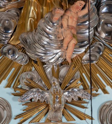 Zdjęcie nr 54: Nastawa architektoniczna, jednoosiowa, jednokondygnacyjna, ustawiona na wysokim cokole. 
Ołtarz drewniany, prostopadłościenny. Na nim tabernakulum z tronem wystawienia. Tabernakulum w formie drewnianej, prostopadłościennej skrzynki, obitej blachą, o wypukłej ściance frontowej, zamkniętej profilowanym gzymsem. Drzwiczki w formie stojącego prostokąta zamkniętego łukiem półkolistym, obwiedzione ramą, z wizerunkiem baranka z krzyżem, leżącego na księdze z pieczęciami, w glorii. Tabernakulum w drewnianej obudowie, wpisane w wysoki cokół, na nim tron wystawienia w formie obrotowej niszy w kształcie stojącego prostokąta, z podwieszoną draperią. Nisza flankowana parą pilastrów, przechodzących w spływy wolutowe, na których siedzą putta. Pilastry dźwigają pełne belkowanie, wyłamane na ich osi, częściowo przerwane przez niszę. Pilastry dekorowane uskrzydlonymi główkami anielskimi oraz snopkiem zboża i kiściami winogron. Cały tron wystawienia dekorowany rocaille'm. 
Nastawa ujęta dwiema parami kolumn o kapitelach kompozytowych, dźwigających pełne belkowanie, wyłamane pomiędzy nimi, przerwane nad polem środkowym. Na belkowaniu fragmenty przerwanego, półkolistego przyczółka, na których leżą putta. Całe pole środkowe wypełnia wnęka w formie stojącego prostokąta zamkniętego łukiem półkolistym, ujęta profilowaną ramą, z figurą Chrystusa Ukrzyżowanego. Nad łukiem wnęki, pomiędzy obłokami, gołębica Ducha Świętego w glorii promienistej. Pomiędzy kolumnami płyciny z uskrzydlonymi główkami anielskimi na obłokach, rocaille oraz figury św.  Jana Ewangelisty i Matki Boskiej na rocaille'owych wspornikach. Kondygnacja ujęta uszami utworzonymi z rocaille'u. Nastawa zwieńczona rzeźbą tronującego Boga Ojca z globem i berłem, pośród obłoków i uskrzydlonych główek anielskich, w glorii promienistej. 
Struktura polichromowana w kolorze błękitnym, kolumny, profile i ornamentyka złocone. 
