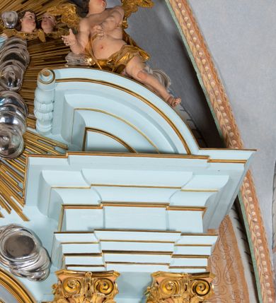 Zdjęcie nr 39: Nastawa architektoniczna, jednoosiowa, jednokondygnacyjna, ustawiona na wysokim cokole. 
Ołtarz drewniany, prostopadłościenny. Na nim tabernakulum z tronem wystawienia. Tabernakulum w formie drewnianej, prostopadłościennej skrzynki, obitej blachą, o wypukłej ściance frontowej, zamkniętej profilowanym gzymsem. Drzwiczki w formie stojącego prostokąta zamkniętego łukiem półkolistym, obwiedzione ramą, z wizerunkiem baranka z krzyżem, leżącego na księdze z pieczęciami, w glorii. Tabernakulum w drewnianej obudowie, wpisane w wysoki cokół, na nim tron wystawienia w formie obrotowej niszy w kształcie stojącego prostokąta, z podwieszoną draperią. Nisza flankowana parą pilastrów, przechodzących w spływy wolutowe, na których siedzą putta. Pilastry dźwigają pełne belkowanie, wyłamane na ich osi, częściowo przerwane przez niszę. Pilastry dekorowane uskrzydlonymi główkami anielskimi oraz snopkiem zboża i kiściami winogron. Cały tron wystawienia dekorowany rocaille'm. 
Nastawa ujęta dwiema parami kolumn o kapitelach kompozytowych, dźwigających pełne belkowanie, wyłamane pomiędzy nimi, przerwane nad polem środkowym. Na belkowaniu fragmenty przerwanego, półkolistego przyczółka, na których leżą putta. Całe pole środkowe wypełnia wnęka w formie stojącego prostokąta zamkniętego łukiem półkolistym, ujęta profilowaną ramą, z figurą Chrystusa Ukrzyżowanego. Nad łukiem wnęki, pomiędzy obłokami, gołębica Ducha Świętego w glorii promienistej. Pomiędzy kolumnami płyciny z uskrzydlonymi główkami anielskimi na obłokach, rocaille oraz figury św.  Jana Ewangelisty i Matki Boskiej na rocaille'owych wspornikach. Kondygnacja ujęta uszami utworzonymi z rocaille'u. Nastawa zwieńczona rzeźbą tronującego Boga Ojca z globem i berłem, pośród obłoków i uskrzydlonych główek anielskich, w glorii promienistej. 
Struktura polichromowana w kolorze błękitnym, kolumny, profile i ornamentyka złocone. 
