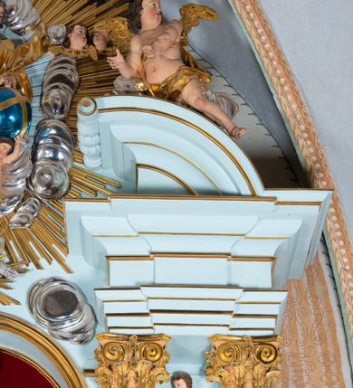 Zdjęcie nr 38: Nastawa architektoniczna, jednoosiowa, jednokondygnacyjna, ustawiona na wysokim cokole. 
Ołtarz drewniany, prostopadłościenny. Na nim tabernakulum z tronem wystawienia. Tabernakulum w formie drewnianej, prostopadłościennej skrzynki, obitej blachą, o wypukłej ściance frontowej, zamkniętej profilowanym gzymsem. Drzwiczki w formie stojącego prostokąta zamkniętego łukiem półkolistym, obwiedzione ramą, z wizerunkiem baranka z krzyżem, leżącego na księdze z pieczęciami, w glorii. Tabernakulum w drewnianej obudowie, wpisane w wysoki cokół, na nim tron wystawienia w formie obrotowej niszy w kształcie stojącego prostokąta, z podwieszoną draperią. Nisza flankowana parą pilastrów, przechodzących w spływy wolutowe, na których siedzą putta. Pilastry dźwigają pełne belkowanie, wyłamane na ich osi, częściowo przerwane przez niszę. Pilastry dekorowane uskrzydlonymi główkami anielskimi oraz snopkiem zboża i kiściami winogron. Cały tron wystawienia dekorowany rocaille&#039;m. 
Nastawa ujęta dwiema parami kolumn o kapitelach kompozytowych, dźwigających pełne belkowanie, wyłamane pomiędzy nimi, przerwane nad polem środkowym. Na belkowaniu fragmenty przerwanego, półkolistego przyczółka, na których leżą putta. Całe pole środkowe wypełnia wnęka w formie stojącego prostokąta zamkniętego łukiem półkolistym, ujęta profilowaną ramą, z figurą Chrystusa Ukrzyżowanego. Nad łukiem wnęki, pomiędzy obłokami, gołębica Ducha Świętego w glorii promienistej. Pomiędzy kolumnami płyciny z uskrzydlonymi główkami anielskimi na obłokach, rocaille oraz figury św.  Jana Ewangelisty i Matki Boskiej na rocaille&#039;owych wspornikach. Kondygnacja ujęta uszami utworzonymi z rocaille&#039;u. Nastawa zwieńczona rzeźbą tronującego Boga Ojca z globem i berłem, pośród obłoków i uskrzydlonych główek anielskich, w glorii promienistej. 
Struktura polichromowana w kolorze błękitnym, kolumny, profile i ornamentyka złocone. 
