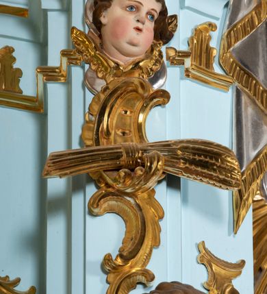 Zdjęcie nr 32: Nastawa architektoniczna, jednoosiowa, jednokondygnacyjna, ustawiona na wysokim cokole. 
Ołtarz drewniany, prostopadłościenny. Na nim tabernakulum z tronem wystawienia. Tabernakulum w formie drewnianej, prostopadłościennej skrzynki, obitej blachą, o wypukłej ściance frontowej, zamkniętej profilowanym gzymsem. Drzwiczki w formie stojącego prostokąta zamkniętego łukiem półkolistym, obwiedzione ramą, z wizerunkiem baranka z krzyżem, leżącego na księdze z pieczęciami, w glorii. Tabernakulum w drewnianej obudowie, wpisane w wysoki cokół, na nim tron wystawienia w formie obrotowej niszy w kształcie stojącego prostokąta, z podwieszoną draperią. Nisza flankowana parą pilastrów, przechodzących w spływy wolutowe, na których siedzą putta. Pilastry dźwigają pełne belkowanie, wyłamane na ich osi, częściowo przerwane przez niszę. Pilastry dekorowane uskrzydlonymi główkami anielskimi oraz snopkiem zboża i kiściami winogron. Cały tron wystawienia dekorowany rocaille'm. 
Nastawa ujęta dwiema parami kolumn o kapitelach kompozytowych, dźwigających pełne belkowanie, wyłamane pomiędzy nimi, przerwane nad polem środkowym. Na belkowaniu fragmenty przerwanego, półkolistego przyczółka, na których leżą putta. Całe pole środkowe wypełnia wnęka w formie stojącego prostokąta zamkniętego łukiem półkolistym, ujęta profilowaną ramą, z figurą Chrystusa Ukrzyżowanego. Nad łukiem wnęki, pomiędzy obłokami, gołębica Ducha Świętego w glorii promienistej. Pomiędzy kolumnami płyciny z uskrzydlonymi główkami anielskimi na obłokach, rocaille oraz figury św.  Jana Ewangelisty i Matki Boskiej na rocaille'owych wspornikach. Kondygnacja ujęta uszami utworzonymi z rocaille'u. Nastawa zwieńczona rzeźbą tronującego Boga Ojca z globem i berłem, pośród obłoków i uskrzydlonych główek anielskich, w glorii promienistej. 
Struktura polichromowana w kolorze błękitnym, kolumny, profile i ornamentyka złocone. 
