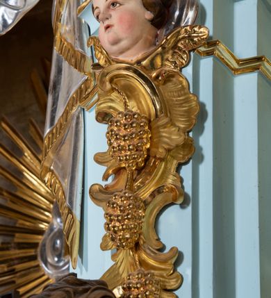 Zdjęcie nr 34: Nastawa architektoniczna, jednoosiowa, jednokondygnacyjna, ustawiona na wysokim cokole. 
Ołtarz drewniany, prostopadłościenny. Na nim tabernakulum z tronem wystawienia. Tabernakulum w formie drewnianej, prostopadłościennej skrzynki, obitej blachą, o wypukłej ściance frontowej, zamkniętej profilowanym gzymsem. Drzwiczki w formie stojącego prostokąta zamkniętego łukiem półkolistym, obwiedzione ramą, z wizerunkiem baranka z krzyżem, leżącego na księdze z pieczęciami, w glorii. Tabernakulum w drewnianej obudowie, wpisane w wysoki cokół, na nim tron wystawienia w formie obrotowej niszy w kształcie stojącego prostokąta, z podwieszoną draperią. Nisza flankowana parą pilastrów, przechodzących w spływy wolutowe, na których siedzą putta. Pilastry dźwigają pełne belkowanie, wyłamane na ich osi, częściowo przerwane przez niszę. Pilastry dekorowane uskrzydlonymi główkami anielskimi oraz snopkiem zboża i kiściami winogron. Cały tron wystawienia dekorowany rocaille&#039;m. 
Nastawa ujęta dwiema parami kolumn o kapitelach kompozytowych, dźwigających pełne belkowanie, wyłamane pomiędzy nimi, przerwane nad polem środkowym. Na belkowaniu fragmenty przerwanego, półkolistego przyczółka, na których leżą putta. Całe pole środkowe wypełnia wnęka w formie stojącego prostokąta zamkniętego łukiem półkolistym, ujęta profilowaną ramą, z figurą Chrystusa Ukrzyżowanego. Nad łukiem wnęki, pomiędzy obłokami, gołębica Ducha Świętego w glorii promienistej. Pomiędzy kolumnami płyciny z uskrzydlonymi główkami anielskimi na obłokach, rocaille oraz figury św.  Jana Ewangelisty i Matki Boskiej na rocaille&#039;owych wspornikach. Kondygnacja ujęta uszami utworzonymi z rocaille&#039;u. Nastawa zwieńczona rzeźbą tronującego Boga Ojca z globem i berłem, pośród obłoków i uskrzydlonych główek anielskich, w glorii promienistej. 
Struktura polichromowana w kolorze błękitnym, kolumny, profile i ornamentyka złocone. 
