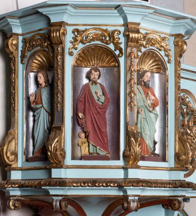 Zdjęcie nr 7: Kosz ambony na planie ośmioboku z szyszką i wolutami u podstawy. Ścianki kosza ujęte pilastrami z karbowaną wstęgą i kampanulami, zakończone od dołu wolutami z motywem liścia akantu oraz wolutą pod kapitelami. W czterech ścianach nisze ze złotymi konchami. W niszach rzeźby czterech ewangelistów z atrybutami. Ponad nimi motywy ornamentalne rocaille z różyczką pośrodku. Na zaplecku tablice Dekalogu na obłoku na tle glorii promienistej. Ujęte w prostokątną ramę zwieńczoną łukiem wklęsło-wypukłym. Baldachim ośmioboczny z podwieszoną glorią i gołębicą, fryz z motywami rocaille&#039;ów i krzyży, wyłamany gzyms. W zwieńczeniu koncentrycznie schodzące się woluty. Ambona polichromowana na kolor seledynowy, detale złocone, postacie w różnobarwnych szatach, partie ciała potraktowane naturalistycznie. 