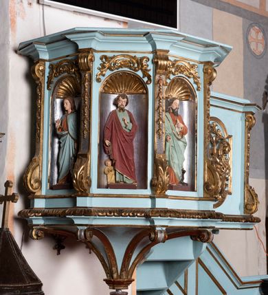 Zdjęcie nr 8: Kosz ambony na planie ośmioboku z szyszką i wolutami u podstawy. Ścianki kosza ujęte pilastrami z karbowaną wstęgą i kampanulami, zakończone od dołu wolutami z motywem liścia akantu oraz wolutą pod kapitelami. W czterech ścianach nisze ze złotymi konchami. W niszach rzeźby czterech ewangelistów z atrybutami. Ponad nimi motywy ornamentalne rocaille z różyczką pośrodku. Na zaplecku tablice Dekalogu na obłoku na tle glorii promienistej. Ujęte w prostokątną ramę zwieńczoną łukiem wklęsło-wypukłym. Baldachim ośmioboczny z podwieszoną glorią i gołębicą, fryz z motywami rocaille'ów i krzyży, wyłamany gzyms. W zwieńczeniu koncentrycznie schodzące się woluty. Ambona polichromowana na kolor seledynowy, detale złocone, postacie w różnobarwnych szatach, partie ciała potraktowane naturalistycznie. 