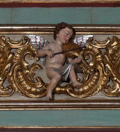 Zdjęcie nr 1: Parapet chóru muzycznego podtrzymywany przez dwie podpory. Balustrada na cokole, podzielona na cztery pola, wydzielone czterema pilastrami o trzonach w formie spływów wolutowych dekorowanych kampanulami i środkową hermą z półpostacią anioła. Pilastry podtrzymują niepełne belkowanie wyłamane na ich osi. Na belkowaniu, na osi skrajnych pilastrów wazony z kwiatami. W polach płyciny w formie leżącego prostokąta, ujęte profilowanymi ramami. W dwóch skrajnych płycinach przedstawienia instrumentów muzycznych ujętych rocaille'm. Dwie środkowe płyciny z figurkami muzykujących puttów flankowanych rocaille'm. Dolna krawędź balustrady, na osi środkowej dekorowana dwiema stykającymi się wolutami, przechodzącymi we wstęgę, ozdobioną rocaille'm. Balustrada polichromowana w kolorze seledynowym, z kolorowymi profilami, ornamentyka i spływy wolutowe złocone i srebrzone. 


