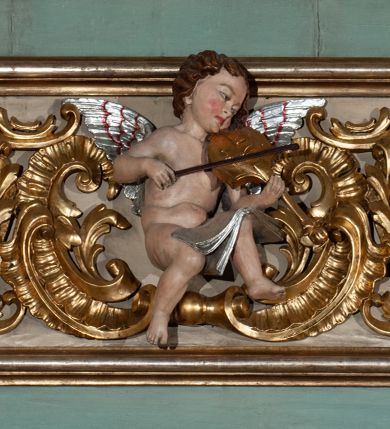 Zdjęcie nr 2: Parapet chóru muzycznego podtrzymywany przez dwie podpory. Balustrada na cokole, podzielona na cztery pola, wydzielone czterema pilastrami o trzonach w formie spływów wolutowych dekorowanych kampanulami i środkową hermą z półpostacią anioła. Pilastry podtrzymują niepełne belkowanie wyłamane na ich osi. Na belkowaniu, na osi skrajnych pilastrów wazony z kwiatami. W polach płyciny w formie leżącego prostokąta, ujęte profilowanymi ramami. W dwóch skrajnych płycinach przedstawienia instrumentów muzycznych ujętych rocaille'm. Dwie środkowe płyciny z figurkami muzykujących puttów flankowanych rocaille'm. Dolna krawędź balustrady, na osi środkowej dekorowana dwiema stykającymi się wolutami, przechodzącymi we wstęgę, ozdobioną rocaille'm. Balustrada polichromowana w kolorze seledynowym, z kolorowymi profilami, ornamentyka i spływy wolutowe złocone i srebrzone. 


