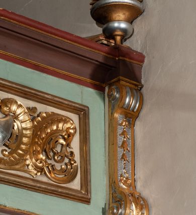 Zdjęcie nr 28: Parapet chóru muzycznego podtrzymywany przez dwie podpory. Balustrada na cokole, podzielona na cztery pola, wydzielone czterema pilastrami o trzonach w formie spływów wolutowych dekorowanych kampanulami i środkową hermą z półpostacią anioła. Pilastry podtrzymują niepełne belkowanie wyłamane na ich osi. Na belkowaniu, na osi skrajnych pilastrów wazony z kwiatami. W polach płyciny w formie leżącego prostokąta, ujęte profilowanymi ramami. W dwóch skrajnych płycinach przedstawienia instrumentów muzycznych ujętych rocaille'm. Dwie środkowe płyciny z figurkami muzykujących puttów flankowanych rocaille'm. Dolna krawędź balustrady, na osi środkowej dekorowana dwiema stykającymi się wolutami, przechodzącymi we wstęgę, ozdobioną rocaille'm. Balustrada polichromowana w kolorze seledynowym, z kolorowymi profilami, ornamentyka i spływy wolutowe złocone i srebrzone. 


