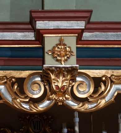 Zdjęcie nr 34: Parapet chóru muzycznego podtrzymywany przez dwie podpory. Balustrada na cokole, podzielona na cztery pola, wydzielone czterema pilastrami o trzonach w formie spływów wolutowych dekorowanych kampanulami i środkową hermą z półpostacią anioła. Pilastry podtrzymują niepełne belkowanie wyłamane na ich osi. Na belkowaniu, na osi skrajnych pilastrów wazony z kwiatami. W polach płyciny w formie leżącego prostokąta, ujęte profilowanymi ramami. W dwóch skrajnych płycinach przedstawienia instrumentów muzycznych ujętych rocaille'm. Dwie środkowe płyciny z figurkami muzykujących puttów flankowanych rocaille'm. Dolna krawędź balustrady, na osi środkowej dekorowana dwiema stykającymi się wolutami, przechodzącymi we wstęgę, ozdobioną rocaille'm. Balustrada polichromowana w kolorze seledynowym, z kolorowymi profilami, ornamentyka i spływy wolutowe złocone i srebrzone. 


