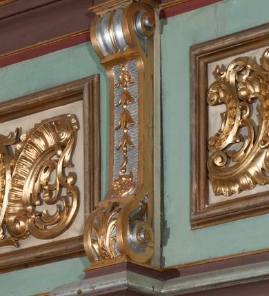 Zdjęcie nr 29: Parapet chóru muzycznego podtrzymywany przez dwie podpory. Balustrada na cokole, podzielona na cztery pola, wydzielone czterema pilastrami o trzonach w formie spływów wolutowych dekorowanych kampanulami i środkową hermą z półpostacią anioła. Pilastry podtrzymują niepełne belkowanie wyłamane na ich osi. Na belkowaniu, na osi skrajnych pilastrów wazony z kwiatami. W polach płyciny w formie leżącego prostokąta, ujęte profilowanymi ramami. W dwóch skrajnych płycinach przedstawienia instrumentów muzycznych ujętych rocaille'm. Dwie środkowe płyciny z figurkami muzykujących puttów flankowanych rocaille'm. Dolna krawędź balustrady, na osi środkowej dekorowana dwiema stykającymi się wolutami, przechodzącymi we wstęgę, ozdobioną rocaille'm. Balustrada polichromowana w kolorze seledynowym, z kolorowymi profilami, ornamentyka i spływy wolutowe złocone i srebrzone. 


