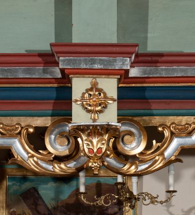 Zdjęcie nr 33: Parapet chóru muzycznego podtrzymywany przez dwie podpory. Balustrada na cokole, podzielona na cztery pola, wydzielone czterema pilastrami o trzonach w formie spływów wolutowych dekorowanych kampanulami i środkową hermą z półpostacią anioła. Pilastry podtrzymują niepełne belkowanie wyłamane na ich osi. Na belkowaniu, na osi skrajnych pilastrów wazony z kwiatami. W polach płyciny w formie leżącego prostokąta, ujęte profilowanymi ramami. W dwóch skrajnych płycinach przedstawienia instrumentów muzycznych ujętych rocaille'm. Dwie środkowe płyciny z figurkami muzykujących puttów flankowanych rocaille'm. Dolna krawędź balustrady, na osi środkowej dekorowana dwiema stykającymi się wolutami, przechodzącymi we wstęgę, ozdobioną rocaille'm. Balustrada polichromowana w kolorze seledynowym, z kolorowymi profilami, ornamentyka i spływy wolutowe złocone i srebrzone. 


