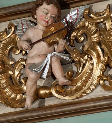 Zdjęcie nr 3: Parapet chóru muzycznego podtrzymywany przez dwie podpory. Balustrada na cokole, podzielona na cztery pola, wydzielone czterema pilastrami o trzonach w formie spływów wolutowych dekorowanych kampanulami i środkową hermą z półpostacią anioła. Pilastry podtrzymują niepełne belkowanie wyłamane na ich osi. Na belkowaniu, na osi skrajnych pilastrów wazony z kwiatami. W polach płyciny w formie leżącego prostokąta, ujęte profilowanymi ramami. W dwóch skrajnych płycinach przedstawienia instrumentów muzycznych ujętych rocaille'm. Dwie środkowe płyciny z figurkami muzykujących puttów flankowanych rocaille'm. Dolna krawędź balustrady, na osi środkowej dekorowana dwiema stykającymi się wolutami, przechodzącymi we wstęgę, ozdobioną rocaille'm. Balustrada polichromowana w kolorze seledynowym, z kolorowymi profilami, ornamentyka i spływy wolutowe złocone i srebrzone. 


