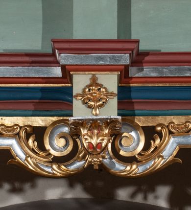Zdjęcie nr 32: Parapet chóru muzycznego podtrzymywany przez dwie podpory. Balustrada na cokole, podzielona na cztery pola, wydzielone czterema pilastrami o trzonach w formie spływów wolutowych dekorowanych kampanulami i środkową hermą z półpostacią anioła. Pilastry podtrzymują niepełne belkowanie wyłamane na ich osi. Na belkowaniu, na osi skrajnych pilastrów wazony z kwiatami. W polach płyciny w formie leżącego prostokąta, ujęte profilowanymi ramami. W dwóch skrajnych płycinach przedstawienia instrumentów muzycznych ujętych rocaille'm. Dwie środkowe płyciny z figurkami muzykujących puttów flankowanych rocaille'm. Dolna krawędź balustrady, na osi środkowej dekorowana dwiema stykającymi się wolutami, przechodzącymi we wstęgę, ozdobioną rocaille'm. Balustrada polichromowana w kolorze seledynowym, z kolorowymi profilami, ornamentyka i spływy wolutowe złocone i srebrzone. 


