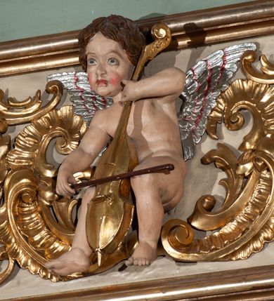Zdjęcie nr 19: Parapet chóru muzycznego podtrzymywany przez dwie podpory. Balustrada na cokole, podzielona na cztery pola, wydzielone czterema pilastrami o trzonach w formie spływów wolutowych dekorowanych kampanulami i środkową hermą z półpostacią anioła. Pilastry podtrzymują niepełne belkowanie wyłamane na ich osi. Na belkowaniu, na osi skrajnych pilastrów wazony z kwiatami. W polach płyciny w formie leżącego prostokąta, ujęte profilowanymi ramami. W dwóch skrajnych płycinach przedstawienia instrumentów muzycznych ujętych rocaille'm. Dwie środkowe płyciny z figurkami muzykujących puttów flankowanych rocaille'm. Dolna krawędź balustrady, na osi środkowej dekorowana dwiema stykającymi się wolutami, przechodzącymi we wstęgę, ozdobioną rocaille'm. Balustrada polichromowana w kolorze seledynowym, z kolorowymi profilami, ornamentyka i spływy wolutowe złocone i srebrzone. 


