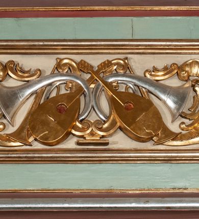 Zdjęcie nr 26: Parapet chóru muzycznego podtrzymywany przez dwie podpory. Balustrada na cokole, podzielona na cztery pola, wydzielone czterema pilastrami o trzonach w formie spływów wolutowych dekorowanych kampanulami i środkową hermą z półpostacią anioła. Pilastry podtrzymują niepełne belkowanie wyłamane na ich osi. Na belkowaniu, na osi skrajnych pilastrów wazony z kwiatami. W polach płyciny w formie leżącego prostokąta, ujęte profilowanymi ramami. W dwóch skrajnych płycinach przedstawienia instrumentów muzycznych ujętych rocaille'm. Dwie środkowe płyciny z figurkami muzykujących puttów flankowanych rocaille'm. Dolna krawędź balustrady, na osi środkowej dekorowana dwiema stykającymi się wolutami, przechodzącymi we wstęgę, ozdobioną rocaille'm. Balustrada polichromowana w kolorze seledynowym, z kolorowymi profilami, ornamentyka i spływy wolutowe złocone i srebrzone. 


