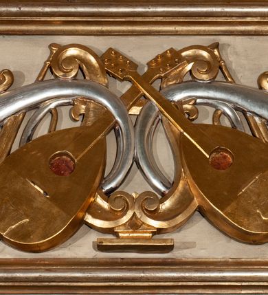 Zdjęcie nr 27: Parapet chóru muzycznego podtrzymywany przez dwie podpory. Balustrada na cokole, podzielona na cztery pola, wydzielone czterema pilastrami o trzonach w formie spływów wolutowych dekorowanych kampanulami i środkową hermą z półpostacią anioła. Pilastry podtrzymują niepełne belkowanie wyłamane na ich osi. Na belkowaniu, na osi skrajnych pilastrów wazony z kwiatami. W polach płyciny w formie leżącego prostokąta, ujęte profilowanymi ramami. W dwóch skrajnych płycinach przedstawienia instrumentów muzycznych ujętych rocaille'm. Dwie środkowe płyciny z figurkami muzykujących puttów flankowanych rocaille'm. Dolna krawędź balustrady, na osi środkowej dekorowana dwiema stykającymi się wolutami, przechodzącymi we wstęgę, ozdobioną rocaille'm. Balustrada polichromowana w kolorze seledynowym, z kolorowymi profilami, ornamentyka i spływy wolutowe złocone i srebrzone. 


