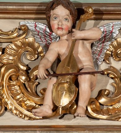 Zdjęcie nr 20: Parapet chóru muzycznego podtrzymywany przez dwie podpory. Balustrada na cokole, podzielona na cztery pola, wydzielone czterema pilastrami o trzonach w formie spływów wolutowych dekorowanych kampanulami i środkową hermą z półpostacią anioła. Pilastry podtrzymują niepełne belkowanie wyłamane na ich osi. Na belkowaniu, na osi skrajnych pilastrów wazony z kwiatami. W polach płyciny w formie leżącego prostokąta, ujęte profilowanymi ramami. W dwóch skrajnych płycinach przedstawienia instrumentów muzycznych ujętych rocaille'm. Dwie środkowe płyciny z figurkami muzykujących puttów flankowanych rocaille'm. Dolna krawędź balustrady, na osi środkowej dekorowana dwiema stykającymi się wolutami, przechodzącymi we wstęgę, ozdobioną rocaille'm. Balustrada polichromowana w kolorze seledynowym, z kolorowymi profilami, ornamentyka i spływy wolutowe złocone i srebrzone. 


