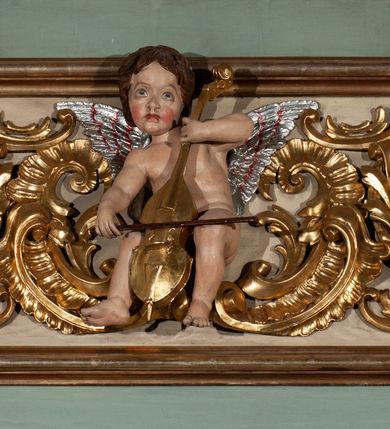 Zdjęcie nr 18: Parapet chóru muzycznego podtrzymywany przez dwie podpory. Balustrada na cokole, podzielona na cztery pola, wydzielone czterema pilastrami o trzonach w formie spływów wolutowych dekorowanych kampanulami i środkową hermą z półpostacią anioła. Pilastry podtrzymują niepełne belkowanie wyłamane na ich osi. Na belkowaniu, na osi skrajnych pilastrów wazony z kwiatami. W polach płyciny w formie leżącego prostokąta, ujęte profilowanymi ramami. W dwóch skrajnych płycinach przedstawienia instrumentów muzycznych ujętych rocaille&#039;m. Dwie środkowe płyciny z figurkami muzykujących puttów flankowanych rocaille&#039;m. Dolna krawędź balustrady, na osi środkowej dekorowana dwiema stykającymi się wolutami, przechodzącymi we wstęgę, ozdobioną rocaille&#039;m. Balustrada polichromowana w kolorze seledynowym, z kolorowymi profilami, ornamentyka i spływy wolutowe złocone i srebrzone. 


