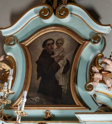 Zdjęcie nr 2: Obraz w formie zbliżonej do stojącego prostokąta zamkniętego łukiem falistym, o wklęsłych bokach. W centrum św. Antoni ukazany do kolan, zwrócony w trzech czwartych w lewo, z uniesioną głową, obiema rękami podtrzymuje Dzieciątko. Twarz ma szczupłą, o dużych, migdałowatych oczach w ciemnej oprawie, skierowanych w górę, prostym, wąskim nosie i pełnych, rozchylonych ustach; okoloną krótkimi, ciemnymi włosami, z tonsurą na głowie i aureolą ponad nią. Ubrany jest w brązowy habit z kapturem, przepasany w talii sznurem, z zawieszonym przy prawym boku różańcem. Dzieciątko w pozycji półsiedzącej, zwrócone w trzech czwartych w prawo, z głową ukazaną frontalnie, obiema rękami obejmuje św. Antoniego. Twarz ma pełną, o dużych, migdałowatych oczach, małym nosie i pełnych ustach; okoloną krótkimi, jasnymi włosami. Wokół głowy ma nimb. Owinięte jest białą tkaniną, odsłaniającą lewe ramię. W lewym dolnym rogu obrazu leżąca gałązka lilii. Postacie ukazane na cieniowanym brązowo-szarym tle. 