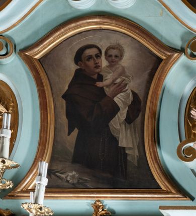Zdjęcie nr 1: Obraz w formie zbliżonej do stojącego prostokąta zamkniętego łukiem falistym, o wklęsłych bokach. W centrum św. Antoni ukazany do kolan, zwrócony w trzech czwartych w lewo, z uniesioną głową, obiema rękami podtrzymuje Dzieciątko. Twarz ma szczupłą, o dużych, migdałowatych oczach w ciemnej oprawie, skierowanych w górę, prostym, wąskim nosie i pełnych, rozchylonych ustach; okoloną krótkimi, ciemnymi włosami, z tonsurą na głowie i aureolą ponad nią. Ubrany jest w brązowy habit z kapturem, przepasany w talii sznurem, z zawieszonym przy prawym boku różańcem. Dzieciątko w pozycji półsiedzącej, zwrócone w trzech czwartych w prawo, z głową ukazaną frontalnie, obiema rękami obejmuje św. Antoniego. Twarz ma pełną, o dużych, migdałowatych oczach, małym nosie i pełnych ustach; okoloną krótkimi, jasnymi włosami. Wokół głowy ma nimb. Owinięte jest białą tkaniną, odsłaniającą lewe ramię. W lewym dolnym rogu obrazu leżąca gałązka lilii. Postacie ukazane na cieniowanym brązowo-szarym tle. 