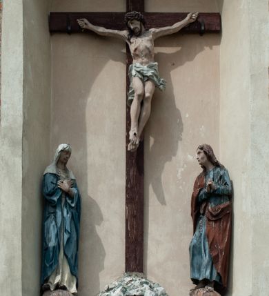 Zdjęcie nr 1: Grupa rzeźbiarska umieszczona we wnęce w formie stojącego prostokąta zamkniętego łukiem półkolistym. W centrum krzyż wypełniający całą wnękę, osadzony na skale, do którego trzema gwoździami przybita jest figura umarłego Chrystusa. Ciało ma w lekkim zwisie, o horyzontalnie rozłożonych ramionach, jego głowa opada na prawy bark, nogi ma lekko ugięte w kolanach, a stopy skrzyżowane w układzie prawa na lewą. Twarz ma szczupłą, o zamkniętych oczach, dużym nosie i otwartych ustach, okoloną krótką, kręconą brodą oraz falowanymi, brązowymi włosami, opadającymi na prawe ramię. Na głowie ma szeroką, brązową koronę cierniową. Ciało o lekko podkreślonej muskulaturze i silnie zaznaczonych żebrach klatki piersiowej. Przez biodra przewiązane krótkie, białe perizonium ze zwisem tkaniny na prawym boku. 
Po lewej stronie krzyża figura Marii, ustawiona na półkolistej podstawie. Matka Boska zwrócona w trzech czwartych w lewo, z pochyloną głową oraz rękami skrzyżowanymi na piersi. Twarz ma owalną, o dużych, migdałowatych oczach, dużym nosie i wąskich ustach. Ubrana jest w białą suknię oraz jasnoniebieski płaszcz z szerokimi rękawami, rozchylony z przodu, na głowę ma narzucony biały welon opadający na ramiona. 
Po prawej stronie krzyża figura św. Jana ustawiona na półkolistej podstawie. Święty zwrócony w trzech czwartych w prawo, w kontrapoście, z uniesionymi, splecionymi dłońmi. Twarz ma szczupłą, o migdałowatych oczach, prostym nosie i wąskich ustach, okoloną gęstymi, kręconymi, brązowymi włosami, opadającymi na ramiona. Ubrany jest w jasnoniebieską suknię oraz zawiązany diagonalnie przez prawe ramię czerwony płaszcz. Polichromia ciał naturalistyczna. 
