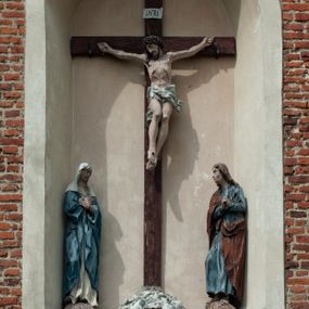 Zdjęcie nr 1: Grupa rzeźbiarska umieszczona we wnęce w formie stojącego prostokąta zamkniętego łukiem półkolistym. W centrum krzyż wypełniający całą wnękę, osadzony na skale, do którego trzema gwoździami przybita jest figura umarłego Chrystusa. Ciało ma w lekkim zwisie, o horyzontalnie rozłożonych ramionach, jego głowa opada na prawy bark, nogi ma lekko ugięte w kolanach, a stopy skrzyżowane w układzie prawa na lewą. Twarz ma szczupłą, o zamkniętych oczach, dużym nosie i otwartych ustach, okoloną krótką, kręconą brodą oraz falowanymi, brązowymi włosami, opadającymi na prawe ramię. Na głowie ma szeroką, brązową koronę cierniową. Ciało o lekko podkreślonej muskulaturze i silnie zaznaczonych żebrach klatki piersiowej. Przez biodra przewiązane krótkie, białe perizonium ze zwisem tkaniny na prawym boku. 
Po lewej stronie krzyża figura Marii, ustawiona na półkolistej podstawie. Matka Boska zwrócona w trzech czwartych w lewo, z pochyloną głową oraz rękami skrzyżowanymi na piersi. Twarz ma owalną, o dużych, migdałowatych oczach, dużym nosie i wąskich ustach. Ubrana jest w białą suknię oraz jasnoniebieski płaszcz z szerokimi rękawami, rozchylony z przodu, na głowę ma narzucony biały welon opadający na ramiona. 
Po prawej stronie krzyża figura św. Jana ustawiona na półkolistej podstawie. Święty zwrócony w trzech czwartych w prawo, w kontrapoście, z uniesionymi, splecionymi dłońmi. Twarz ma szczupłą, o migdałowatych oczach, prostym nosie i wąskich ustach, okoloną gęstymi, kręconymi, brązowymi włosami, opadającymi na ramiona. Ubrany jest w jasnoniebieską suknię oraz zawiązany diagonalnie przez prawe ramię czerwony płaszcz. Polichromia ciał naturalistyczna. 
