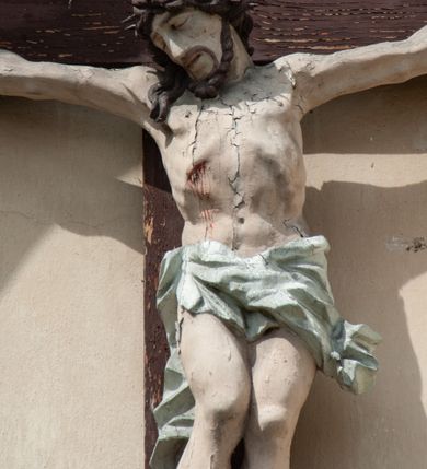 Zdjęcie nr 4: Grupa rzeźbiarska umieszczona we wnęce w formie stojącego prostokąta zamkniętego łukiem półkolistym. W centrum krzyż wypełniający całą wnękę, osadzony na skale, do którego trzema gwoździami przybita jest figura umarłego Chrystusa. Ciało ma w lekkim zwisie, o horyzontalnie rozłożonych ramionach, jego głowa opada na prawy bark, nogi ma lekko ugięte w kolanach, a stopy skrzyżowane w układzie prawa na lewą. Twarz ma szczupłą, o zamkniętych oczach, dużym nosie i otwartych ustach, okoloną krótką, kręconą brodą oraz falowanymi, brązowymi włosami, opadającymi na prawe ramię. Na głowie ma szeroką, brązową koronę cierniową. Ciało o lekko podkreślonej muskulaturze i silnie zaznaczonych żebrach klatki piersiowej. Przez biodra przewiązane krótkie, białe perizonium ze zwisem tkaniny na prawym boku. 
Po lewej stronie krzyża figura Marii, ustawiona na półkolistej podstawie. Matka Boska zwrócona w trzech czwartych w lewo, z pochyloną głową oraz rękami skrzyżowanymi na piersi. Twarz ma owalną, o dużych, migdałowatych oczach, dużym nosie i wąskich ustach. Ubrana jest w białą suknię oraz jasnoniebieski płaszcz z szerokimi rękawami, rozchylony z przodu, na głowę ma narzucony biały welon opadający na ramiona. 
Po prawej stronie krzyża figura św. Jana ustawiona na półkolistej podstawie. Święty zwrócony w trzech czwartych w prawo, w kontrapoście, z uniesionymi, splecionymi dłońmi. Twarz ma szczupłą, o migdałowatych oczach, prostym nosie i wąskich ustach, okoloną gęstymi, kręconymi, brązowymi włosami, opadającymi na ramiona. Ubrany jest w jasnoniebieską suknię oraz zawiązany diagonalnie przez prawe ramię czerwony płaszcz. Polichromia ciał naturalistyczna. 
