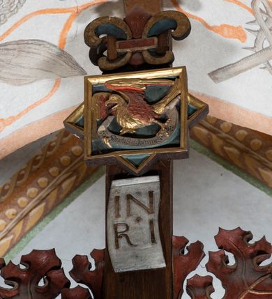 Zdjęcie nr 9: Rzeźba pełnoplastyczna, z tyłu opracowana. Ciało Chrystusa przybite do krzyża na niemal horyzontalnie, szeroko rozłożonych ramionach. Głowa w koronie cierniowej pochylona na prawe ramię. Długie włosy w puklach o spiralnie zakończonych lokach opadają na ramiona i plecy. Twarz szczupła o zindywidualizowanych rysach, pełna spokoju i smutku, okolona krótkim zarostem, z wydatnymi kośćmi policzkowymi, z kroplami krwi na czole. Duże, okrągłe oczy przesłonięte powiekami, podkreślone uniesionymi łukami brwiowymi. Usta drobne, zaciśnięte, nos prosty, wąski. Ręce chude długie, silnie wyprężone, dłonie rozchylone, przebite gwoździami. Klatka piersiowa o starannie opracowanej anatomii, z silnym wcięciem w talii oraz raną w prawym boku. Perizonium w kształcie chusty, sięgające do połowy uda, miękko udrapowane z przodu, spływające po obu bokach kaskadowymi fałdami. Nogi wyprostowane, umięśnione, ułożone obok siebie, stopy stosunkowo duże, skrzyżowane, założone prawa na lewą, przebite jednym gwoździem. Z ran na boku, dłoniach i stopach spływa obficie krew. Karnacja Chrystusa jasnoróżowa, włosy brązowe z domalowanym warkoczem z tyłu, korona zielona, perizonium złocone, z niebieską podszewką. Krzyż łaciński, dębowy, w naturalnym kolorze drewna, wykonany w 1890, w formie Drzewa Życia, z rozłożystymi liśćmi uschniętego akantu w kolorze czerwonym i złotym żyłkowaniem na skrzyżowaniu ramion. W zakończeniu ramion lilijki czerwono-zielone ze złoconymi obwódkami oraz nakładane plakiety w układzie koncentrycznym tworzące ośmioramienne gwiazdy z płaskorzeźbionymi symbolami Ewangelistów. Tła plakiet malowane na niebiesko, symbole i obramienia złocone. Plakiety współczesne z rzeźbą Chrystusa, gotyckie - 1433 r.; u góry krzyża - symbol św. Jana - orzeł, u dołu - symbol św. Łukasza - wół, na prawym ramieniu symbol św. Mateusza - anioł, na lewym ramieniu - symbol św. Marka - lew. Ponad głową Chrystusa titulus z napisem: INRI. Plakieta kwadratowa nałożona na romb w układzie koncentrycznym, w profilowanym obramieniu, z płaskorzeźbionym przedstawieniem orła; u dołu banderola z napisem minuskułą gotycką: sanctus joannes. Plakieta kwadratowa nałożona na romb w układzie koncentrycznym, w obramieniu profilowanym, z fazą wklęsłą - z płaskorzeźbionym przedstawieniem anioła w pozycji siedzącej z podkurczonymi nogami w ujęciu frontalnym, w rękach podtrzymującego faliście wygiętą banderolę o zrolowanych zakończeniach, z czarnym napisem minuskułą gotycką: sanctus matheus; banderola biała z czarnymi rozetkami. Twarz anioła okrągła, o rysach dziecka, różowa, okolona puklami rudych włosów. Anioł ubrany w fałdzistą szatę, złoconą z trójkątnym dekoltem, udrapowaną na kolanach w głębokie, łyżkowate fałdy. U ramion szeroko rozpostarte skrzydła o schematycznych rombowych podziałach, zagięte na brzegach, zakończone długimi piórami, lotkami - złocone. Tło lekko pogłębione, niebieskie; obramienia kwadratów złocone, boki i tylne ściany malowane na brązowo. Plakieta kwadratowa nałożona na romb w układzie koncentrycznym, w obramieniu profilowanym, z fazą wklęsłą, z płaskorzeźbionym przedstawieniem uskrzydlonego wołu, w pełnej postaci, w ujęciu bocznym, o masywnej budowie ciała, w dynamicznym ruchu, wspartego na rozstawionych nogach, zakończonych ciężkimi kopytami, z puszystym ogonem. Głowa zwrócona na wprost, oczy duże, nozdrza rozdęte, uszy odstające na boki, pośrodku ozdobnie osadzone esowato wygięte rogi. Skrzydła wyrastające z barków, uniesione w górę, rozpostarte, upierzenie schematyczne zaznaczone w kształcie rombów, z długimi lotkami w zakończeniach wygiętych skrzydeł. U dołu, pomiędzy nogami zwierzęcia - półkoliście wygięta biała banderola, z czarnym napisem minuskułą gotycką: sanctus lucas i czarnymi rozetkami. Tło nieco pogłębione, niebieskie. Postać wołu, obramienie plakiet, złocone; ścianki tylne malowane na brązowo. Plakieta w kształcie rombu nałożonego na kwadrat w układzie koncentrycznym, w obramieniu profilowanym, z fazą wklęsłą - z płaskorzeźbionym przedstawieniem uskrzydlonego lwa. W pełnej postaci, ujęciu bocznym, muskularnych kształtach, mocnych nogach z potężnymi pazurami. Głowa zwrócona na wprost, ujęta bujną, silnie skręconą grzywą. Skrzydła wyrastające z barków uniesione w górę, szeroko rozpostarte, zakończone długimi lotkami; upierzenie schematycznie zaznaczone w formie regularnych rombów. Ogon cienki, długi, z puszystą kitką, przechodzący pomiędzy nogami, esowato wygięty nad grzbietem. U dołu biała banderola, podtrzymywana pazurami przednich łap, z napisem czarnym, gotycką minuskułą: sanctus marcus i czarnymi rozetkami. Tło nieco pogłębione, niebieskie; postać lwa i obramienia złocone, tylne ścianki malowane na brązowo.