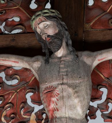 Zdjęcie nr 8: Rzeźba pełnoplastyczna, z tyłu opracowana. Ciało Chrystusa przybite do krzyża na niemal horyzontalnie, szeroko rozłożonych ramionach. Głowa w koronie cierniowej pochylona na prawe ramię. Długie włosy w puklach o spiralnie zakończonych lokach opadają na ramiona i plecy. Twarz szczupła o zindywidualizowanych rysach, pełna spokoju i smutku, okolona krótkim zarostem, z wydatnymi kośćmi policzkowymi, z kroplami krwi na czole. Duże, okrągłe oczy przesłonięte powiekami, podkreślone uniesionymi łukami brwiowymi. Usta drobne, zaciśnięte, nos prosty, wąski. Ręce chude długie, silnie wyprężone, dłonie rozchylone, przebite gwoździami. Klatka piersiowa o starannie opracowanej anatomii, z silnym wcięciem w talii oraz raną w prawym boku. Perizonium w kształcie chusty, sięgające do połowy uda, miękko udrapowane z przodu, spływające po obu bokach kaskadowymi fałdami. Nogi wyprostowane, umięśnione, ułożone obok siebie, stopy stosunkowo duże, skrzyżowane, założone prawa na lewą, przebite jednym gwoździem. Z ran na boku, dłoniach i stopach spływa obficie krew. Karnacja Chrystusa jasnoróżowa, włosy brązowe z domalowanym warkoczem z tyłu, korona zielona, perizonium złocone, z niebieską podszewką. Krzyż łaciński, dębowy, w naturalnym kolorze drewna, wykonany w 1890, w formie Drzewa Życia, z rozłożystymi liśćmi uschniętego akantu w kolorze czerwonym i złotym żyłkowaniem na skrzyżowaniu ramion. W zakończeniu ramion lilijki czerwono-zielone ze złoconymi obwódkami oraz nakładane plakiety w układzie koncentrycznym tworzące ośmioramienne gwiazdy z płaskorzeźbionymi symbolami Ewangelistów. Tła plakiet malowane na niebiesko, symbole i obramienia złocone. Plakiety współczesne z rzeźbą Chrystusa, gotyckie - 1433 r.; u góry krzyża - symbol św. Jana - orzeł, u dołu - symbol św. Łukasza - wół, na prawym ramieniu symbol św. Mateusza - anioł, na lewym ramieniu - symbol św. Marka - lew. Ponad głową Chrystusa titulus z napisem: INRI. Plakieta kwadratowa nałożona na romb w układzie koncentrycznym, w profilowanym obramieniu, z płaskorzeźbionym przedstawieniem orła; u dołu banderola z napisem minuskułą gotycką: sanctus joannes. Plakieta kwadratowa nałożona na romb w układzie koncentrycznym, w obramieniu profilowanym, z fazą wklęsłą - z płaskorzeźbionym przedstawieniem anioła w pozycji siedzącej z podkurczonymi nogami w ujęciu frontalnym, w rękach podtrzymującego faliście wygiętą banderolę o zrolowanych zakończeniach, z czarnym napisem minuskułą gotycką: sanctus matheus; banderola biała z czarnymi rozetkami. Twarz anioła okrągła, o rysach dziecka, różowa, okolona puklami rudych włosów. Anioł ubrany w fałdzistą szatę, złoconą z trójkątnym dekoltem, udrapowaną na kolanach w głębokie, łyżkowate fałdy. U ramion szeroko rozpostarte skrzydła o schematycznych rombowych podziałach, zagięte na brzegach, zakończone długimi piórami, lotkami - złocone. Tło lekko pogłębione, niebieskie; obramienia kwadratów złocone, boki i tylne ściany malowane na brązowo. Plakieta kwadratowa nałożona na romb w układzie koncentrycznym, w obramieniu profilowanym, z fazą wklęsłą, z płaskorzeźbionym przedstawieniem uskrzydlonego wołu, w pełnej postaci, w ujęciu bocznym, o masywnej budowie ciała, w dynamicznym ruchu, wspartego na rozstawionych nogach, zakończonych ciężkimi kopytami, z puszystym ogonem. Głowa zwrócona na wprost, oczy duże, nozdrza rozdęte, uszy odstające na boki, pośrodku ozdobnie osadzone esowato wygięte rogi. Skrzydła wyrastające z barków, uniesione w górę, rozpostarte, upierzenie schematyczne zaznaczone w kształcie rombów, z długimi lotkami w zakończeniach wygiętych skrzydeł. U dołu, pomiędzy nogami zwierzęcia - półkoliście wygięta biała banderola, z czarnym napisem minuskułą gotycką: sanctus lucas i czarnymi rozetkami. Tło nieco pogłębione, niebieskie. Postać wołu, obramienie plakiet, złocone; ścianki tylne malowane na brązowo. Plakieta w kształcie rombu nałożonego na kwadrat w układzie koncentrycznym, w obramieniu profilowanym, z fazą wklęsłą - z płaskorzeźbionym przedstawieniem uskrzydlonego lwa. W pełnej postaci, ujęciu bocznym, muskularnych kształtach, mocnych nogach z potężnymi pazurami. Głowa zwrócona na wprost, ujęta bujną, silnie skręconą grzywą. Skrzydła wyrastające z barków uniesione w górę, szeroko rozpostarte, zakończone długimi lotkami; upierzenie schematycznie zaznaczone w formie regularnych rombów. Ogon cienki, długi, z puszystą kitką, przechodzący pomiędzy nogami, esowato wygięty nad grzbietem. U dołu biała banderola, podtrzymywana pazurami przednich łap, z napisem czarnym, gotycką minuskułą: sanctus marcus i czarnymi rozetkami. Tło nieco pogłębione, niebieskie; postać lwa i obramienia złocone, tylne ścianki malowane na brązowo.
