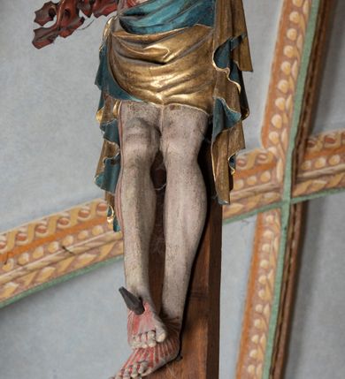 Zdjęcie nr 10: Rzeźba pełnoplastyczna, z tyłu opracowana. Ciało Chrystusa przybite do krzyża na niemal horyzontalnie, szeroko rozłożonych ramionach. Głowa w koronie cierniowej pochylona na prawe ramię. Długie włosy w puklach o spiralnie zakończonych lokach opadają na ramiona i plecy. Twarz szczupła o zindywidualizowanych rysach, pełna spokoju i smutku, okolona krótkim zarostem, z wydatnymi kośćmi policzkowymi, z kroplami krwi na czole. Duże, okrągłe oczy przesłonięte powiekami, podkreślone uniesionymi łukami brwiowymi. Usta drobne, zaciśnięte, nos prosty, wąski. Ręce chude długie, silnie wyprężone, dłonie rozchylone, przebite gwoździami. Klatka piersiowa o starannie opracowanej anatomii, z silnym wcięciem w talii oraz raną w prawym boku. Perizonium w kształcie chusty, sięgające do połowy uda, miękko udrapowane z przodu, spływające po obu bokach kaskadowymi fałdami. Nogi wyprostowane, umięśnione, ułożone obok siebie, stopy stosunkowo duże, skrzyżowane, założone prawa na lewą, przebite jednym gwoździem. Z ran na boku, dłoniach i stopach spływa obficie krew. Karnacja Chrystusa jasnoróżowa, włosy brązowe z domalowanym warkoczem z tyłu, korona zielona, perizonium złocone, z niebieską podszewką. Krzyż łaciński, dębowy, w naturalnym kolorze drewna, wykonany w 1890, w formie Drzewa Życia, z rozłożystymi liśćmi uschniętego akantu w kolorze czerwonym i złotym żyłkowaniem na skrzyżowaniu ramion. W zakończeniu ramion lilijki czerwono-zielone ze złoconymi obwódkami oraz nakładane plakiety w układzie koncentrycznym tworzące ośmioramienne gwiazdy z płaskorzeźbionymi symbolami Ewangelistów. Tła plakiet malowane na niebiesko, symbole i obramienia złocone. Plakiety współczesne z rzeźbą Chrystusa, gotyckie - 1433 r.; u góry krzyża - symbol św. Jana - orzeł, u dołu - symbol św. Łukasza - wół, na prawym ramieniu symbol św. Mateusza - anioł, na lewym ramieniu - symbol św. Marka - lew. Ponad głową Chrystusa titulus z napisem: INRI. Plakieta kwadratowa nałożona na romb w układzie koncentrycznym, w profilowanym obramieniu, z płaskorzeźbionym przedstawieniem orła; u dołu banderola z napisem minuskułą gotycką: sanctus joannes. Plakieta kwadratowa nałożona na romb w układzie koncentrycznym, w obramieniu profilowanym, z fazą wklęsłą - z płaskorzeźbionym przedstawieniem anioła w pozycji siedzącej z podkurczonymi nogami w ujęciu frontalnym, w rękach podtrzymującego faliście wygiętą banderolę o zrolowanych zakończeniach, z czarnym napisem minuskułą gotycką: sanctus matheus; banderola biała z czarnymi rozetkami. Twarz anioła okrągła, o rysach dziecka, różowa, okolona puklami rudych włosów. Anioł ubrany w fałdzistą szatę, złoconą z trójkątnym dekoltem, udrapowaną na kolanach w głębokie, łyżkowate fałdy. U ramion szeroko rozpostarte skrzydła o schematycznych rombowych podziałach, zagięte na brzegach, zakończone długimi piórami, lotkami - złocone. Tło lekko pogłębione, niebieskie; obramienia kwadratów złocone, boki i tylne ściany malowane na brązowo. Plakieta kwadratowa nałożona na romb w układzie koncentrycznym, w obramieniu profilowanym, z fazą wklęsłą, z płaskorzeźbionym przedstawieniem uskrzydlonego wołu, w pełnej postaci, w ujęciu bocznym, o masywnej budowie ciała, w dynamicznym ruchu, wspartego na rozstawionych nogach, zakończonych ciężkimi kopytami, z puszystym ogonem. Głowa zwrócona na wprost, oczy duże, nozdrza rozdęte, uszy odstające na boki, pośrodku ozdobnie osadzone esowato wygięte rogi. Skrzydła wyrastające z barków, uniesione w górę, rozpostarte, upierzenie schematyczne zaznaczone w kształcie rombów, z długimi lotkami w zakończeniach wygiętych skrzydeł. U dołu, pomiędzy nogami zwierzęcia - półkoliście wygięta biała banderola, z czarnym napisem minuskułą gotycką: sanctus lucas i czarnymi rozetkami. Tło nieco pogłębione, niebieskie. Postać wołu, obramienie plakiet, złocone; ścianki tylne malowane na brązowo. Plakieta w kształcie rombu nałożonego na kwadrat w układzie koncentrycznym, w obramieniu profilowanym, z fazą wklęsłą - z płaskorzeźbionym przedstawieniem uskrzydlonego lwa. W pełnej postaci, ujęciu bocznym, muskularnych kształtach, mocnych nogach z potężnymi pazurami. Głowa zwrócona na wprost, ujęta bujną, silnie skręconą grzywą. Skrzydła wyrastające z barków uniesione w górę, szeroko rozpostarte, zakończone długimi lotkami; upierzenie schematycznie zaznaczone w formie regularnych rombów. Ogon cienki, długi, z puszystą kitką, przechodzący pomiędzy nogami, esowato wygięty nad grzbietem. U dołu biała banderola, podtrzymywana pazurami przednich łap, z napisem czarnym, gotycką minuskułą: sanctus marcus i czarnymi rozetkami. Tło nieco pogłębione, niebieskie; postać lwa i obramienia złocone, tylne ścianki malowane na brązowo.