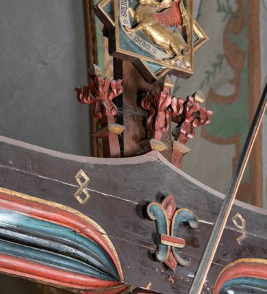 Zdjęcie nr 11: Rzeźba pełnoplastyczna, z tyłu opracowana. Ciało Chrystusa przybite do krzyża na niemal horyzontalnie, szeroko rozłożonych ramionach. Głowa w koronie cierniowej pochylona na prawe ramię. Długie włosy w puklach o spiralnie zakończonych lokach opadają na ramiona i plecy. Twarz szczupła o zindywidualizowanych rysach, pełna spokoju i smutku, okolona krótkim zarostem, z wydatnymi kośćmi policzkowymi, z kroplami krwi na czole. Duże, okrągłe oczy przesłonięte powiekami, podkreślone uniesionymi łukami brwiowymi. Usta drobne, zaciśnięte, nos prosty, wąski. Ręce chude długie, silnie wyprężone, dłonie rozchylone, przebite gwoździami. Klatka piersiowa o starannie opracowanej anatomii, z silnym wcięciem w talii oraz raną w prawym boku. Perizonium w kształcie chusty, sięgające do połowy uda, miękko udrapowane z przodu, spływające po obu bokach kaskadowymi fałdami. Nogi wyprostowane, umięśnione, ułożone obok siebie, stopy stosunkowo duże, skrzyżowane, założone prawa na lewą, przebite jednym gwoździem. Z ran na boku, dłoniach i stopach spływa obficie krew. Karnacja Chrystusa jasnoróżowa, włosy brązowe z domalowanym warkoczem z tyłu, korona zielona, perizonium złocone, z niebieską podszewką. Krzyż łaciński, dębowy, w naturalnym kolorze drewna, wykonany w 1890, w formie Drzewa Życia, z rozłożystymi liśćmi uschniętego akantu w kolorze czerwonym i złotym żyłkowaniem na skrzyżowaniu ramion. W zakończeniu ramion lilijki czerwono-zielone ze złoconymi obwódkami oraz nakładane plakiety w układzie koncentrycznym tworzące ośmioramienne gwiazdy z płaskorzeźbionymi symbolami Ewangelistów. Tła plakiet malowane na niebiesko, symbole i obramienia złocone. Plakiety współczesne z rzeźbą Chrystusa, gotyckie - 1433 r.; u góry krzyża - symbol św. Jana - orzeł, u dołu - symbol św. Łukasza - wół, na prawym ramieniu symbol św. Mateusza - anioł, na lewym ramieniu - symbol św. Marka - lew. Ponad głową Chrystusa titulus z napisem: INRI. Plakieta kwadratowa nałożona na romb w układzie koncentrycznym, w profilowanym obramieniu, z płaskorzeźbionym przedstawieniem orła; u dołu banderola z napisem minuskułą gotycką: sanctus joannes. Plakieta kwadratowa nałożona na romb w układzie koncentrycznym, w obramieniu profilowanym, z fazą wklęsłą - z płaskorzeźbionym przedstawieniem anioła w pozycji siedzącej z podkurczonymi nogami w ujęciu frontalnym, w rękach podtrzymującego faliście wygiętą banderolę o zrolowanych zakończeniach, z czarnym napisem minuskułą gotycką: sanctus matheus; banderola biała z czarnymi rozetkami. Twarz anioła okrągła, o rysach dziecka, różowa, okolona puklami rudych włosów. Anioł ubrany w fałdzistą szatę, złoconą z trójkątnym dekoltem, udrapowaną na kolanach w głębokie, łyżkowate fałdy. U ramion szeroko rozpostarte skrzydła o schematycznych rombowych podziałach, zagięte na brzegach, zakończone długimi piórami, lotkami - złocone. Tło lekko pogłębione, niebieskie; obramienia kwadratów złocone, boki i tylne ściany malowane na brązowo. Plakieta kwadratowa nałożona na romb w układzie koncentrycznym, w obramieniu profilowanym, z fazą wklęsłą, z płaskorzeźbionym przedstawieniem uskrzydlonego wołu, w pełnej postaci, w ujęciu bocznym, o masywnej budowie ciała, w dynamicznym ruchu, wspartego na rozstawionych nogach, zakończonych ciężkimi kopytami, z puszystym ogonem. Głowa zwrócona na wprost, oczy duże, nozdrza rozdęte, uszy odstające na boki, pośrodku ozdobnie osadzone esowato wygięte rogi. Skrzydła wyrastające z barków, uniesione w górę, rozpostarte, upierzenie schematyczne zaznaczone w kształcie rombów, z długimi lotkami w zakończeniach wygiętych skrzydeł. U dołu, pomiędzy nogami zwierzęcia - półkoliście wygięta biała banderola, z czarnym napisem minuskułą gotycką: sanctus lucas i czarnymi rozetkami. Tło nieco pogłębione, niebieskie. Postać wołu, obramienie plakiet, złocone; ścianki tylne malowane na brązowo. Plakieta w kształcie rombu nałożonego na kwadrat w układzie koncentrycznym, w obramieniu profilowanym, z fazą wklęsłą - z płaskorzeźbionym przedstawieniem uskrzydlonego lwa. W pełnej postaci, ujęciu bocznym, muskularnych kształtach, mocnych nogach z potężnymi pazurami. Głowa zwrócona na wprost, ujęta bujną, silnie skręconą grzywą. Skrzydła wyrastające z barków uniesione w górę, szeroko rozpostarte, zakończone długimi lotkami; upierzenie schematycznie zaznaczone w formie regularnych rombów. Ogon cienki, długi, z puszystą kitką, przechodzący pomiędzy nogami, esowato wygięty nad grzbietem. U dołu biała banderola, podtrzymywana pazurami przednich łap, z napisem czarnym, gotycką minuskułą: sanctus marcus i czarnymi rozetkami. Tło nieco pogłębione, niebieskie; postać lwa i obramienia złocone, tylne ścianki malowane na brązowo.