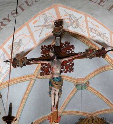 Zdjęcie nr 1: Rzeźba pełnoplastyczna, z tyłu opracowana. Ciało Chrystusa przybite do krzyża na niemal horyzontalnie, szeroko rozłożonych ramionach. Głowa w koronie cierniowej pochylona na prawe ramię. Długie włosy w puklach o spiralnie zakończonych lokach opadają na ramiona i plecy. Twarz szczupła o zindywidualizowanych rysach, pełna spokoju i smutku, okolona krótkim zarostem, z wydatnymi kośćmi policzkowymi, z kroplami krwi na czole. Duże, okrągłe oczy przesłonięte powiekami, podkreślone uniesionymi łukami brwiowymi. Usta drobne, zaciśnięte, nos prosty, wąski. Ręce chude długie, silnie wyprężone, dłonie rozchylone, przebite gwoździami. Klatka piersiowa o starannie opracowanej anatomii, z silnym wcięciem w talii oraz raną w prawym boku. Perizonium w kształcie chusty, sięgające do połowy uda, miękko udrapowane z przodu, spływające po obu bokach kaskadowymi fałdami. Nogi wyprostowane, umięśnione, ułożone obok siebie, stopy stosunkowo duże, skrzyżowane, założone prawa na lewą, przebite jednym gwoździem. Z ran na boku, dłoniach i stopach spływa obficie krew. Karnacja Chrystusa jasnoróżowa, włosy brązowe z domalowanym warkoczem z tyłu, korona zielona, perizonium złocone, z niebieską podszewką. Krzyż łaciński, dębowy, w naturalnym kolorze drewna, wykonany w 1890, w formie Drzewa Życia, z rozłożystymi liśćmi uschniętego akantu w kolorze czerwonym i złotym żyłkowaniem na skrzyżowaniu ramion. W zakończeniu ramion lilijki czerwono-zielone ze złoconymi obwódkami oraz nakładane plakiety w układzie koncentrycznym tworzące ośmioramienne gwiazdy z płaskorzeźbionymi symbolami Ewangelistów. Tła plakiet malowane na niebiesko, symbole i obramienia złocone. Plakiety współczesne z rzeźbą Chrystusa, gotyckie - 1433 r.; u góry krzyża - symbol św. Jana - orzeł, u dołu - symbol św. Łukasza - wół, na prawym ramieniu symbol św. Mateusza - anioł, na lewym ramieniu - symbol św. Marka - lew. Ponad głową Chrystusa titulus z napisem: INRI. Plakieta kwadratowa nałożona na romb w układzie koncentrycznym, w profilowanym obramieniu, z płaskorzeźbionym przedstawieniem orła; u dołu banderola z napisem minuskułą gotycką: sanctus joannes. Plakieta kwadratowa nałożona na romb w układzie koncentrycznym, w obramieniu profilowanym, z fazą wklęsłą - z płaskorzeźbionym przedstawieniem anioła w pozycji siedzącej z podkurczonymi nogami w ujęciu frontalnym, w rękach podtrzymującego faliście wygiętą banderolę o zrolowanych zakończeniach, z czarnym napisem minuskułą gotycką: sanctus matheus; banderola biała z czarnymi rozetkami. Twarz anioła okrągła, o rysach dziecka, różowa, okolona puklami rudych włosów. Anioł ubrany w fałdzistą szatę, złoconą z trójkątnym dekoltem, udrapowaną na kolanach w głębokie, łyżkowate fałdy. U ramion szeroko rozpostarte skrzydła o schematycznych rombowych podziałach, zagięte na brzegach, zakończone długimi piórami, lotkami - złocone. Tło lekko pogłębione, niebieskie; obramienia kwadratów złocone, boki i tylne ściany malowane na brązowo. Plakieta kwadratowa nałożona na romb w układzie koncentrycznym, w obramieniu profilowanym, z fazą wklęsłą, z płaskorzeźbionym przedstawieniem uskrzydlonego wołu, w pełnej postaci, w ujęciu bocznym, o masywnej budowie ciała, w dynamicznym ruchu, wspartego na rozstawionych nogach, zakończonych ciężkimi kopytami, z puszystym ogonem. Głowa zwrócona na wprost, oczy duże, nozdrza rozdęte, uszy odstające na boki, pośrodku ozdobnie osadzone esowato wygięte rogi. Skrzydła wyrastające z barków, uniesione w górę, rozpostarte, upierzenie schematyczne zaznaczone w kształcie rombów, z długimi lotkami w zakończeniach wygiętych skrzydeł. U dołu, pomiędzy nogami zwierzęcia - półkoliście wygięta biała banderola, z czarnym napisem minuskułą gotycką: sanctus lucas i czarnymi rozetkami. Tło nieco pogłębione, niebieskie. Postać wołu, obramienie plakiet, złocone; ścianki tylne malowane na brązowo. Plakieta w kształcie rombu nałożonego na kwadrat w układzie koncentrycznym, w obramieniu profilowanym, z fazą wklęsłą - z płaskorzeźbionym przedstawieniem uskrzydlonego lwa. W pełnej postaci, ujęciu bocznym, muskularnych kształtach, mocnych nogach z potężnymi pazurami. Głowa zwrócona na wprost, ujęta bujną, silnie skręconą grzywą. Skrzydła wyrastające z barków uniesione w górę, szeroko rozpostarte, zakończone długimi lotkami; upierzenie schematycznie zaznaczone w formie regularnych rombów. Ogon cienki, długi, z puszystą kitką, przechodzący pomiędzy nogami, esowato wygięty nad grzbietem. U dołu biała banderola, podtrzymywana pazurami przednich łap, z napisem czarnym, gotycką minuskułą: sanctus marcus i czarnymi rozetkami. Tło nieco pogłębione, niebieskie; postać lwa i obramienia złocone, tylne ścianki malowane na brązowo.