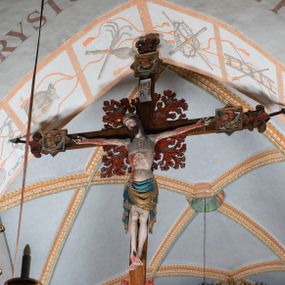 Zdjęcie nr 1: Rzeźba pełnoplastyczna, z tyłu opracowana. Ciało Chrystusa przybite do krzyża na niemal horyzontalnie, szeroko rozłożonych ramionach. Głowa w koronie cierniowej pochylona na prawe ramię. Długie włosy w puklach o spiralnie zakończonych lokach opadają na ramiona i plecy. Twarz szczupła o zindywidualizowanych rysach, pełna spokoju i smutku, okolona krótkim zarostem, z wydatnymi kośćmi policzkowymi, z kroplami krwi na czole. Duże, okrągłe oczy przesłonięte powiekami, podkreślone uniesionymi łukami brwiowymi. Usta drobne, zaciśnięte, nos prosty, wąski. Ręce chude długie, silnie wyprężone, dłonie rozchylone, przebite gwoździami. Klatka piersiowa o starannie opracowanej anatomii, z silnym wcięciem w talii oraz raną w prawym boku. Perizonium w kształcie chusty, sięgające do połowy uda, miękko udrapowane z przodu, spływające po obu bokach kaskadowymi fałdami. Nogi wyprostowane, umięśnione, ułożone obok siebie, stopy stosunkowo duże, skrzyżowane, założone prawa na lewą, przebite jednym gwoździem. Z ran na boku, dłoniach i stopach spływa obficie krew. Karnacja Chrystusa jasnoróżowa, włosy brązowe z domalowanym warkoczem z tyłu, korona zielona, perizonium złocone, z niebieską podszewką. Krzyż łaciński, dębowy, w naturalnym kolorze drewna, wykonany w 1890, w formie Drzewa Życia, z rozłożystymi liśćmi uschniętego akantu w kolorze czerwonym i złotym żyłkowaniem na skrzyżowaniu ramion. W zakończeniu ramion lilijki czerwono-zielone ze złoconymi obwódkami oraz nakładane plakiety w układzie koncentrycznym tworzące ośmioramienne gwiazdy z płaskorzeźbionymi symbolami Ewangelistów. Tła plakiet malowane na niebiesko, symbole i obramienia złocone. Plakiety współczesne z rzeźbą Chrystusa, gotyckie - 1433 r.; u góry krzyża - symbol św. Jana - orzeł, u dołu - symbol św. Łukasza - wół, na prawym ramieniu symbol św. Mateusza - anioł, na lewym ramieniu - symbol św. Marka - lew. Ponad głową Chrystusa titulus z napisem: INRI. Plakieta kwadratowa nałożona na romb w układzie koncentrycznym, w profilowanym obramieniu, z płaskorzeźbionym przedstawieniem orła; u dołu banderola z napisem minuskułą gotycką: sanctus joannes. Plakieta kwadratowa nałożona na romb w układzie koncentrycznym, w obramieniu profilowanym, z fazą wklęsłą - z płaskorzeźbionym przedstawieniem anioła w pozycji siedzącej z podkurczonymi nogami w ujęciu frontalnym, w rękach podtrzymującego faliście wygiętą banderolę o zrolowanych zakończeniach, z czarnym napisem minuskułą gotycką: sanctus matheus; banderola biała z czarnymi rozetkami. Twarz anioła okrągła, o rysach dziecka, różowa, okolona puklami rudych włosów. Anioł ubrany w fałdzistą szatę, złoconą z trójkątnym dekoltem, udrapowaną na kolanach w głębokie, łyżkowate fałdy. U ramion szeroko rozpostarte skrzydła o schematycznych rombowych podziałach, zagięte na brzegach, zakończone długimi piórami, lotkami - złocone. Tło lekko pogłębione, niebieskie; obramienia kwadratów złocone, boki i tylne ściany malowane na brązowo. Plakieta kwadratowa nałożona na romb w układzie koncentrycznym, w obramieniu profilowanym, z fazą wklęsłą, z płaskorzeźbionym przedstawieniem uskrzydlonego wołu, w pełnej postaci, w ujęciu bocznym, o masywnej budowie ciała, w dynamicznym ruchu, wspartego na rozstawionych nogach, zakończonych ciężkimi kopytami, z puszystym ogonem. Głowa zwrócona na wprost, oczy duże, nozdrza rozdęte, uszy odstające na boki, pośrodku ozdobnie osadzone esowato wygięte rogi. Skrzydła wyrastające z barków, uniesione w górę, rozpostarte, upierzenie schematyczne zaznaczone w kształcie rombów, z długimi lotkami w zakończeniach wygiętych skrzydeł. U dołu, pomiędzy nogami zwierzęcia - półkoliście wygięta biała banderola, z czarnym napisem minuskułą gotycką: sanctus lucas i czarnymi rozetkami. Tło nieco pogłębione, niebieskie. Postać wołu, obramienie plakiet, złocone; ścianki tylne malowane na brązowo. Plakieta w kształcie rombu nałożonego na kwadrat w układzie koncentrycznym, w obramieniu profilowanym, z fazą wklęsłą - z płaskorzeźbionym przedstawieniem uskrzydlonego lwa. W pełnej postaci, ujęciu bocznym, muskularnych kształtach, mocnych nogach z potężnymi pazurami. Głowa zwrócona na wprost, ujęta bujną, silnie skręconą grzywą. Skrzydła wyrastające z barków uniesione w górę, szeroko rozpostarte, zakończone długimi lotkami; upierzenie schematycznie zaznaczone w formie regularnych rombów. Ogon cienki, długi, z puszystą kitką, przechodzący pomiędzy nogami, esowato wygięty nad grzbietem. U dołu biała banderola, podtrzymywana pazurami przednich łap, z napisem czarnym, gotycką minuskułą: sanctus marcus i czarnymi rozetkami. Tło nieco pogłębione, niebieskie; postać lwa i obramienia złocone, tylne ścianki malowane na brązowo.