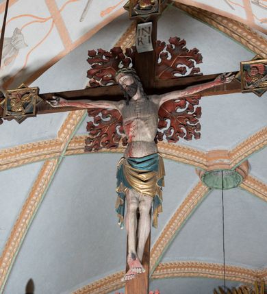 Zdjęcie nr 14: Rzeźba pełnoplastyczna, z tyłu opracowana. Ciało Chrystusa przybite do krzyża na niemal horyzontalnie, szeroko rozłożonych ramionach. Głowa w koronie cierniowej pochylona na prawe ramię. Długie włosy w puklach o spiralnie zakończonych lokach opadają na ramiona i plecy. Twarz szczupła o zindywidualizowanych rysach, pełna spokoju i smutku, okolona krótkim zarostem, z wydatnymi kośćmi policzkowymi, z kroplami krwi na czole. Duże, okrągłe oczy przesłonięte powiekami, podkreślone uniesionymi łukami brwiowymi. Usta drobne, zaciśnięte, nos prosty, wąski. Ręce chude długie, silnie wyprężone, dłonie rozchylone, przebite gwoździami. Klatka piersiowa o starannie opracowanej anatomii, z silnym wcięciem w talii oraz raną w prawym boku. Perizonium w kształcie chusty, sięgające do połowy uda, miękko udrapowane z przodu, spływające po obu bokach kaskadowymi fałdami. Nogi wyprostowane, umięśnione, ułożone obok siebie, stopy stosunkowo duże, skrzyżowane, założone prawa na lewą, przebite jednym gwoździem. Z ran na boku, dłoniach i stopach spływa obficie krew. Karnacja Chrystusa jasnoróżowa, włosy brązowe z domalowanym warkoczem z tyłu, korona zielona, perizonium złocone, z niebieską podszewką. Krzyż łaciński, dębowy, w naturalnym kolorze drewna, wykonany w 1890, w formie Drzewa Życia, z rozłożystymi liśćmi uschniętego akantu w kolorze czerwonym i złotym żyłkowaniem na skrzyżowaniu ramion. W zakończeniu ramion lilijki czerwono-zielone ze złoconymi obwódkami oraz nakładane plakiety w układzie koncentrycznym tworzące ośmioramienne gwiazdy z płaskorzeźbionymi symbolami Ewangelistów. Tła plakiet malowane na niebiesko, symbole i obramienia złocone. Plakiety współczesne z rzeźbą Chrystusa, gotyckie - 1433 r.; u góry krzyża - symbol św. Jana - orzeł, u dołu - symbol św. Łukasza - wół, na prawym ramieniu symbol św. Mateusza - anioł, na lewym ramieniu - symbol św. Marka - lew. Ponad głową Chrystusa titulus z napisem: INRI. Plakieta kwadratowa nałożona na romb w układzie koncentrycznym, w profilowanym obramieniu, z płaskorzeźbionym przedstawieniem orła; u dołu banderola z napisem minuskułą gotycką: sanctus joannes. Plakieta kwadratowa nałożona na romb w układzie koncentrycznym, w obramieniu profilowanym, z fazą wklęsłą - z płaskorzeźbionym przedstawieniem anioła w pozycji siedzącej z podkurczonymi nogami w ujęciu frontalnym, w rękach podtrzymującego faliście wygiętą banderolę o zrolowanych zakończeniach, z czarnym napisem minuskułą gotycką: sanctus matheus; banderola biała z czarnymi rozetkami. Twarz anioła okrągła, o rysach dziecka, różowa, okolona puklami rudych włosów. Anioł ubrany w fałdzistą szatę, złoconą z trójkątnym dekoltem, udrapowaną na kolanach w głębokie, łyżkowate fałdy. U ramion szeroko rozpostarte skrzydła o schematycznych rombowych podziałach, zagięte na brzegach, zakończone długimi piórami, lotkami - złocone. Tło lekko pogłębione, niebieskie; obramienia kwadratów złocone, boki i tylne ściany malowane na brązowo. Plakieta kwadratowa nałożona na romb w układzie koncentrycznym, w obramieniu profilowanym, z fazą wklęsłą, z płaskorzeźbionym przedstawieniem uskrzydlonego wołu, w pełnej postaci, w ujęciu bocznym, o masywnej budowie ciała, w dynamicznym ruchu, wspartego na rozstawionych nogach, zakończonych ciężkimi kopytami, z puszystym ogonem. Głowa zwrócona na wprost, oczy duże, nozdrza rozdęte, uszy odstające na boki, pośrodku ozdobnie osadzone esowato wygięte rogi. Skrzydła wyrastające z barków, uniesione w górę, rozpostarte, upierzenie schematyczne zaznaczone w kształcie rombów, z długimi lotkami w zakończeniach wygiętych skrzydeł. U dołu, pomiędzy nogami zwierzęcia - półkoliście wygięta biała banderola, z czarnym napisem minuskułą gotycką: sanctus lucas i czarnymi rozetkami. Tło nieco pogłębione, niebieskie. Postać wołu, obramienie plakiet, złocone; ścianki tylne malowane na brązowo. Plakieta w kształcie rombu nałożonego na kwadrat w układzie koncentrycznym, w obramieniu profilowanym, z fazą wklęsłą - z płaskorzeźbionym przedstawieniem uskrzydlonego lwa. W pełnej postaci, ujęciu bocznym, muskularnych kształtach, mocnych nogach z potężnymi pazurami. Głowa zwrócona na wprost, ujęta bujną, silnie skręconą grzywą. Skrzydła wyrastające z barków uniesione w górę, szeroko rozpostarte, zakończone długimi lotkami; upierzenie schematycznie zaznaczone w formie regularnych rombów. Ogon cienki, długi, z puszystą kitką, przechodzący pomiędzy nogami, esowato wygięty nad grzbietem. U dołu biała banderola, podtrzymywana pazurami przednich łap, z napisem czarnym, gotycką minuskułą: sanctus marcus i czarnymi rozetkami. Tło nieco pogłębione, niebieskie; postać lwa i obramienia złocone, tylne ścianki malowane na brązowo.