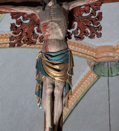 Zdjęcie nr 15: Rzeźba pełnoplastyczna, z tyłu opracowana. Ciało Chrystusa przybite do krzyża na niemal horyzontalnie, szeroko rozłożonych ramionach. Głowa w koronie cierniowej pochylona na prawe ramię. Długie włosy w puklach o spiralnie zakończonych lokach opadają na ramiona i plecy. Twarz szczupła o zindywidualizowanych rysach, pełna spokoju i smutku, okolona krótkim zarostem, z wydatnymi kośćmi policzkowymi, z kroplami krwi na czole. Duże, okrągłe oczy przesłonięte powiekami, podkreślone uniesionymi łukami brwiowymi. Usta drobne, zaciśnięte, nos prosty, wąski. Ręce chude długie, silnie wyprężone, dłonie rozchylone, przebite gwoździami. Klatka piersiowa o starannie opracowanej anatomii, z silnym wcięciem w talii oraz raną w prawym boku. Perizonium w kształcie chusty, sięgające do połowy uda, miękko udrapowane z przodu, spływające po obu bokach kaskadowymi fałdami. Nogi wyprostowane, umięśnione, ułożone obok siebie, stopy stosunkowo duże, skrzyżowane, założone prawa na lewą, przebite jednym gwoździem. Z ran na boku, dłoniach i stopach spływa obficie krew. Karnacja Chrystusa jasnoróżowa, włosy brązowe z domalowanym warkoczem z tyłu, korona zielona, perizonium złocone, z niebieską podszewką. Krzyż łaciński, dębowy, w naturalnym kolorze drewna, wykonany w 1890, w formie Drzewa Życia, z rozłożystymi liśćmi uschniętego akantu w kolorze czerwonym i złotym żyłkowaniem na skrzyżowaniu ramion. W zakończeniu ramion lilijki czerwono-zielone ze złoconymi obwódkami oraz nakładane plakiety w układzie koncentrycznym tworzące ośmioramienne gwiazdy z płaskorzeźbionymi symbolami Ewangelistów. Tła plakiet malowane na niebiesko, symbole i obramienia złocone. Plakiety współczesne z rzeźbą Chrystusa, gotyckie - 1433 r.; u góry krzyża - symbol św. Jana - orzeł, u dołu - symbol św. Łukasza - wół, na prawym ramieniu symbol św. Mateusza - anioł, na lewym ramieniu - symbol św. Marka - lew. Ponad głową Chrystusa titulus z napisem: INRI. Plakieta kwadratowa nałożona na romb w układzie koncentrycznym, w profilowanym obramieniu, z płaskorzeźbionym przedstawieniem orła; u dołu banderola z napisem minuskułą gotycką: sanctus joannes. Plakieta kwadratowa nałożona na romb w układzie koncentrycznym, w obramieniu profilowanym, z fazą wklęsłą - z płaskorzeźbionym przedstawieniem anioła w pozycji siedzącej z podkurczonymi nogami w ujęciu frontalnym, w rękach podtrzymującego faliście wygiętą banderolę o zrolowanych zakończeniach, z czarnym napisem minuskułą gotycką: sanctus matheus; banderola biała z czarnymi rozetkami. Twarz anioła okrągła, o rysach dziecka, różowa, okolona puklami rudych włosów. Anioł ubrany w fałdzistą szatę, złoconą z trójkątnym dekoltem, udrapowaną na kolanach w głębokie, łyżkowate fałdy. U ramion szeroko rozpostarte skrzydła o schematycznych rombowych podziałach, zagięte na brzegach, zakończone długimi piórami, lotkami - złocone. Tło lekko pogłębione, niebieskie; obramienia kwadratów złocone, boki i tylne ściany malowane na brązowo. Plakieta kwadratowa nałożona na romb w układzie koncentrycznym, w obramieniu profilowanym, z fazą wklęsłą, z płaskorzeźbionym przedstawieniem uskrzydlonego wołu, w pełnej postaci, w ujęciu bocznym, o masywnej budowie ciała, w dynamicznym ruchu, wspartego na rozstawionych nogach, zakończonych ciężkimi kopytami, z puszystym ogonem. Głowa zwrócona na wprost, oczy duże, nozdrza rozdęte, uszy odstające na boki, pośrodku ozdobnie osadzone esowato wygięte rogi. Skrzydła wyrastające z barków, uniesione w górę, rozpostarte, upierzenie schematyczne zaznaczone w kształcie rombów, z długimi lotkami w zakończeniach wygiętych skrzydeł. U dołu, pomiędzy nogami zwierzęcia - półkoliście wygięta biała banderola, z czarnym napisem minuskułą gotycką: sanctus lucas i czarnymi rozetkami. Tło nieco pogłębione, niebieskie. Postać wołu, obramienie plakiet, złocone; ścianki tylne malowane na brązowo. Plakieta w kształcie rombu nałożonego na kwadrat w układzie koncentrycznym, w obramieniu profilowanym, z fazą wklęsłą - z płaskorzeźbionym przedstawieniem uskrzydlonego lwa. W pełnej postaci, ujęciu bocznym, muskularnych kształtach, mocnych nogach z potężnymi pazurami. Głowa zwrócona na wprost, ujęta bujną, silnie skręconą grzywą. Skrzydła wyrastające z barków uniesione w górę, szeroko rozpostarte, zakończone długimi lotkami; upierzenie schematycznie zaznaczone w formie regularnych rombów. Ogon cienki, długi, z puszystą kitką, przechodzący pomiędzy nogami, esowato wygięty nad grzbietem. U dołu biała banderola, podtrzymywana pazurami przednich łap, z napisem czarnym, gotycką minuskułą: sanctus marcus i czarnymi rozetkami. Tło nieco pogłębione, niebieskie; postać lwa i obramienia złocone, tylne ścianki malowane na brązowo.