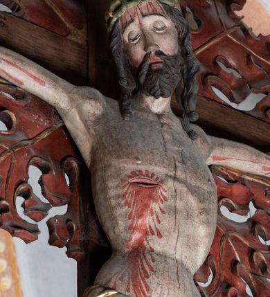 Zdjęcie nr 12: Rzeźba pełnoplastyczna, z tyłu opracowana. Ciało Chrystusa przybite do krzyża na niemal horyzontalnie, szeroko rozłożonych ramionach. Głowa w koronie cierniowej pochylona na prawe ramię. Długie włosy w puklach o spiralnie zakończonych lokach opadają na ramiona i plecy. Twarz szczupła o zindywidualizowanych rysach, pełna spokoju i smutku, okolona krótkim zarostem, z wydatnymi kośćmi policzkowymi, z kroplami krwi na czole. Duże, okrągłe oczy przesłonięte powiekami, podkreślone uniesionymi łukami brwiowymi. Usta drobne, zaciśnięte, nos prosty, wąski. Ręce chude długie, silnie wyprężone, dłonie rozchylone, przebite gwoździami. Klatka piersiowa o starannie opracowanej anatomii, z silnym wcięciem w talii oraz raną w prawym boku. Perizonium w kształcie chusty, sięgające do połowy uda, miękko udrapowane z przodu, spływające po obu bokach kaskadowymi fałdami. Nogi wyprostowane, umięśnione, ułożone obok siebie, stopy stosunkowo duże, skrzyżowane, założone prawa na lewą, przebite jednym gwoździem. Z ran na boku, dłoniach i stopach spływa obficie krew. Karnacja Chrystusa jasnoróżowa, włosy brązowe z domalowanym warkoczem z tyłu, korona zielona, perizonium złocone, z niebieską podszewką. Krzyż łaciński, dębowy, w naturalnym kolorze drewna, wykonany w 1890, w formie Drzewa Życia, z rozłożystymi liśćmi uschniętego akantu w kolorze czerwonym i złotym żyłkowaniem na skrzyżowaniu ramion. W zakończeniu ramion lilijki czerwono-zielone ze złoconymi obwódkami oraz nakładane plakiety w układzie koncentrycznym tworzące ośmioramienne gwiazdy z płaskorzeźbionymi symbolami Ewangelistów. Tła plakiet malowane na niebiesko, symbole i obramienia złocone. Plakiety współczesne z rzeźbą Chrystusa, gotyckie - 1433 r.; u góry krzyża - symbol św. Jana - orzeł, u dołu - symbol św. Łukasza - wół, na prawym ramieniu symbol św. Mateusza - anioł, na lewym ramieniu - symbol św. Marka - lew. Ponad głową Chrystusa titulus z napisem: INRI. Plakieta kwadratowa nałożona na romb w układzie koncentrycznym, w profilowanym obramieniu, z płaskorzeźbionym przedstawieniem orła; u dołu banderola z napisem minuskułą gotycką: sanctus joannes. Plakieta kwadratowa nałożona na romb w układzie koncentrycznym, w obramieniu profilowanym, z fazą wklęsłą - z płaskorzeźbionym przedstawieniem anioła w pozycji siedzącej z podkurczonymi nogami w ujęciu frontalnym, w rękach podtrzymującego faliście wygiętą banderolę o zrolowanych zakończeniach, z czarnym napisem minuskułą gotycką: sanctus matheus; banderola biała z czarnymi rozetkami. Twarz anioła okrągła, o rysach dziecka, różowa, okolona puklami rudych włosów. Anioł ubrany w fałdzistą szatę, złoconą z trójkątnym dekoltem, udrapowaną na kolanach w głębokie, łyżkowate fałdy. U ramion szeroko rozpostarte skrzydła o schematycznych rombowych podziałach, zagięte na brzegach, zakończone długimi piórami, lotkami - złocone. Tło lekko pogłębione, niebieskie; obramienia kwadratów złocone, boki i tylne ściany malowane na brązowo. Plakieta kwadratowa nałożona na romb w układzie koncentrycznym, w obramieniu profilowanym, z fazą wklęsłą, z płaskorzeźbionym przedstawieniem uskrzydlonego wołu, w pełnej postaci, w ujęciu bocznym, o masywnej budowie ciała, w dynamicznym ruchu, wspartego na rozstawionych nogach, zakończonych ciężkimi kopytami, z puszystym ogonem. Głowa zwrócona na wprost, oczy duże, nozdrza rozdęte, uszy odstające na boki, pośrodku ozdobnie osadzone esowato wygięte rogi. Skrzydła wyrastające z barków, uniesione w górę, rozpostarte, upierzenie schematyczne zaznaczone w kształcie rombów, z długimi lotkami w zakończeniach wygiętych skrzydeł. U dołu, pomiędzy nogami zwierzęcia - półkoliście wygięta biała banderola, z czarnym napisem minuskułą gotycką: sanctus lucas i czarnymi rozetkami. Tło nieco pogłębione, niebieskie. Postać wołu, obramienie plakiet, złocone; ścianki tylne malowane na brązowo. Plakieta w kształcie rombu nałożonego na kwadrat w układzie koncentrycznym, w obramieniu profilowanym, z fazą wklęsłą - z płaskorzeźbionym przedstawieniem uskrzydlonego lwa. W pełnej postaci, ujęciu bocznym, muskularnych kształtach, mocnych nogach z potężnymi pazurami. Głowa zwrócona na wprost, ujęta bujną, silnie skręconą grzywą. Skrzydła wyrastające z barków uniesione w górę, szeroko rozpostarte, zakończone długimi lotkami; upierzenie schematycznie zaznaczone w formie regularnych rombów. Ogon cienki, długi, z puszystą kitką, przechodzący pomiędzy nogami, esowato wygięty nad grzbietem. U dołu biała banderola, podtrzymywana pazurami przednich łap, z napisem czarnym, gotycką minuskułą: sanctus marcus i czarnymi rozetkami. Tło nieco pogłębione, niebieskie; postać lwa i obramienia złocone, tylne ścianki malowane na brązowo.