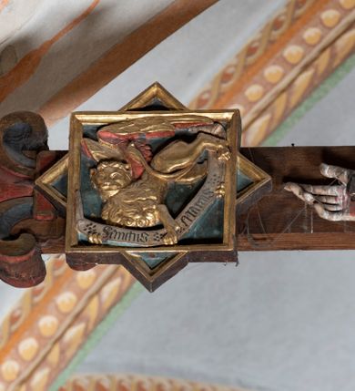 Zdjęcie nr 6: Rzeźba pełnoplastyczna, z tyłu opracowana. Ciało Chrystusa przybite do krzyża na niemal horyzontalnie, szeroko rozłożonych ramionach. Głowa w koronie cierniowej pochylona na prawe ramię. Długie włosy w puklach o spiralnie zakończonych lokach opadają na ramiona i plecy. Twarz szczupła o zindywidualizowanych rysach, pełna spokoju i smutku, okolona krótkim zarostem, z wydatnymi kośćmi policzkowymi, z kroplami krwi na czole. Duże, okrągłe oczy przesłonięte powiekami, podkreślone uniesionymi łukami brwiowymi. Usta drobne, zaciśnięte, nos prosty, wąski. Ręce chude długie, silnie wyprężone, dłonie rozchylone, przebite gwoździami. Klatka piersiowa o starannie opracowanej anatomii, z silnym wcięciem w talii oraz raną w prawym boku. Perizonium w kształcie chusty, sięgające do połowy uda, miękko udrapowane z przodu, spływające po obu bokach kaskadowymi fałdami. Nogi wyprostowane, umięśnione, ułożone obok siebie, stopy stosunkowo duże, skrzyżowane, założone prawa na lewą, przebite jednym gwoździem. Z ran na boku, dłoniach i stopach spływa obficie krew. Karnacja Chrystusa jasnoróżowa, włosy brązowe z domalowanym warkoczem z tyłu, korona zielona, perizonium złocone, z niebieską podszewką. Krzyż łaciński, dębowy, w naturalnym kolorze drewna, wykonany w 1890, w formie Drzewa Życia, z rozłożystymi liśćmi uschniętego akantu w kolorze czerwonym i złotym żyłkowaniem na skrzyżowaniu ramion. W zakończeniu ramion lilijki czerwono-zielone ze złoconymi obwódkami oraz nakładane plakiety w układzie koncentrycznym tworzące ośmioramienne gwiazdy z płaskorzeźbionymi symbolami Ewangelistów. Tła plakiet malowane na niebiesko, symbole i obramienia złocone. Plakiety współczesne z rzeźbą Chrystusa, gotyckie - 1433 r.; u góry krzyża - symbol św. Jana - orzeł, u dołu - symbol św. Łukasza - wół, na prawym ramieniu symbol św. Mateusza - anioł, na lewym ramieniu - symbol św. Marka - lew. Ponad głową Chrystusa titulus z napisem: INRI. Plakieta kwadratowa nałożona na romb w układzie koncentrycznym, w profilowanym obramieniu, z płaskorzeźbionym przedstawieniem orła; u dołu banderola z napisem minuskułą gotycką: sanctus joannes. Plakieta kwadratowa nałożona na romb w układzie koncentrycznym, w obramieniu profilowanym, z fazą wklęsłą - z płaskorzeźbionym przedstawieniem anioła w pozycji siedzącej z podkurczonymi nogami w ujęciu frontalnym, w rękach podtrzymującego faliście wygiętą banderolę o zrolowanych zakończeniach, z czarnym napisem minuskułą gotycką: sanctus matheus; banderola biała z czarnymi rozetkami. Twarz anioła okrągła, o rysach dziecka, różowa, okolona puklami rudych włosów. Anioł ubrany w fałdzistą szatę, złoconą z trójkątnym dekoltem, udrapowaną na kolanach w głębokie, łyżkowate fałdy. U ramion szeroko rozpostarte skrzydła o schematycznych rombowych podziałach, zagięte na brzegach, zakończone długimi piórami, lotkami - złocone. Tło lekko pogłębione, niebieskie; obramienia kwadratów złocone, boki i tylne ściany malowane na brązowo. Plakieta kwadratowa nałożona na romb w układzie koncentrycznym, w obramieniu profilowanym, z fazą wklęsłą, z płaskorzeźbionym przedstawieniem uskrzydlonego wołu, w pełnej postaci, w ujęciu bocznym, o masywnej budowie ciała, w dynamicznym ruchu, wspartego na rozstawionych nogach, zakończonych ciężkimi kopytami, z puszystym ogonem. Głowa zwrócona na wprost, oczy duże, nozdrza rozdęte, uszy odstające na boki, pośrodku ozdobnie osadzone esowato wygięte rogi. Skrzydła wyrastające z barków, uniesione w górę, rozpostarte, upierzenie schematyczne zaznaczone w kształcie rombów, z długimi lotkami w zakończeniach wygiętych skrzydeł. U dołu, pomiędzy nogami zwierzęcia - półkoliście wygięta biała banderola, z czarnym napisem minuskułą gotycką: sanctus lucas i czarnymi rozetkami. Tło nieco pogłębione, niebieskie. Postać wołu, obramienie plakiet, złocone; ścianki tylne malowane na brązowo. Plakieta w kształcie rombu nałożonego na kwadrat w układzie koncentrycznym, w obramieniu profilowanym, z fazą wklęsłą - z płaskorzeźbionym przedstawieniem uskrzydlonego lwa. W pełnej postaci, ujęciu bocznym, muskularnych kształtach, mocnych nogach z potężnymi pazurami. Głowa zwrócona na wprost, ujęta bujną, silnie skręconą grzywą. Skrzydła wyrastające z barków uniesione w górę, szeroko rozpostarte, zakończone długimi lotkami; upierzenie schematycznie zaznaczone w formie regularnych rombów. Ogon cienki, długi, z puszystą kitką, przechodzący pomiędzy nogami, esowato wygięty nad grzbietem. U dołu biała banderola, podtrzymywana pazurami przednich łap, z napisem czarnym, gotycką minuskułą: sanctus marcus i czarnymi rozetkami. Tło nieco pogłębione, niebieskie; postać lwa i obramienia złocone, tylne ścianki malowane na brązowo.