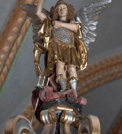 Zdjęcie nr 5: Rzeźba pełnoplastyczna, z tyłu opracowana. Ciało Chrystusa przybite do krzyża na niemal horyzontalnie, szeroko rozłożonych ramionach. Głowa w koronie cierniowej pochylona na prawe ramię. Długie włosy w puklach o spiralnie zakończonych lokach opadają na ramiona i plecy. Twarz szczupła o zindywidualizowanych rysach, pełna spokoju i smutku, okolona krótkim zarostem, z wydatnymi kośćmi policzkowymi, z kroplami krwi na czole. Duże, okrągłe oczy przesłonięte powiekami, podkreślone uniesionymi łukami brwiowymi. Usta drobne, zaciśnięte, nos prosty, wąski. Ręce chude długie, silnie wyprężone, dłonie rozchylone, przebite gwoździami. Klatka piersiowa o starannie opracowanej anatomii, z silnym wcięciem w talii oraz raną w prawym boku. Perizonium w kształcie chusty, sięgające do połowy uda, miękko udrapowane z przodu, spływające po obu bokach kaskadowymi fałdami. Nogi wyprostowane, umięśnione, ułożone obok siebie, stopy stosunkowo duże, skrzyżowane, założone prawa na lewą, przebite jednym gwoździem. Z ran na boku, dłoniach i stopach spływa obficie krew. Karnacja Chrystusa jasnoróżowa, włosy brązowe z domalowanym warkoczem z tyłu, korona zielona, perizonium złocone, z niebieską podszewką. Krzyż łaciński, dębowy, w naturalnym kolorze drewna, wykonany w 1890, w formie Drzewa Życia, z rozłożystymi liśćmi uschniętego akantu w kolorze czerwonym i złotym żyłkowaniem na skrzyżowaniu ramion. W zakończeniu ramion lilijki czerwono-zielone ze złoconymi obwódkami oraz nakładane plakiety w układzie koncentrycznym tworzące ośmioramienne gwiazdy z płaskorzeźbionymi symbolami Ewangelistów. Tła plakiet malowane na niebiesko, symbole i obramienia złocone. Plakiety współczesne z rzeźbą Chrystusa, gotyckie - 1433 r.; u góry krzyża - symbol św. Jana - orzeł, u dołu - symbol św. Łukasza - wół, na prawym ramieniu symbol św. Mateusza - anioł, na lewym ramieniu - symbol św. Marka - lew. Ponad głową Chrystusa titulus z napisem: INRI. Plakieta kwadratowa nałożona na romb w układzie koncentrycznym, w profilowanym obramieniu, z płaskorzeźbionym przedstawieniem orła; u dołu banderola z napisem minuskułą gotycką: sanctus joannes. Plakieta kwadratowa nałożona na romb w układzie koncentrycznym, w obramieniu profilowanym, z fazą wklęsłą - z płaskorzeźbionym przedstawieniem anioła w pozycji siedzącej z podkurczonymi nogami w ujęciu frontalnym, w rękach podtrzymującego faliście wygiętą banderolę o zrolowanych zakończeniach, z czarnym napisem minuskułą gotycką: sanctus matheus; banderola biała z czarnymi rozetkami. Twarz anioła okrągła, o rysach dziecka, różowa, okolona puklami rudych włosów. Anioł ubrany w fałdzistą szatę, złoconą z trójkątnym dekoltem, udrapowaną na kolanach w głębokie, łyżkowate fałdy. U ramion szeroko rozpostarte skrzydła o schematycznych rombowych podziałach, zagięte na brzegach, zakończone długimi piórami, lotkami - złocone. Tło lekko pogłębione, niebieskie; obramienia kwadratów złocone, boki i tylne ściany malowane na brązowo. Plakieta kwadratowa nałożona na romb w układzie koncentrycznym, w obramieniu profilowanym, z fazą wklęsłą, z płaskorzeźbionym przedstawieniem uskrzydlonego wołu, w pełnej postaci, w ujęciu bocznym, o masywnej budowie ciała, w dynamicznym ruchu, wspartego na rozstawionych nogach, zakończonych ciężkimi kopytami, z puszystym ogonem. Głowa zwrócona na wprost, oczy duże, nozdrza rozdęte, uszy odstające na boki, pośrodku ozdobnie osadzone esowato wygięte rogi. Skrzydła wyrastające z barków, uniesione w górę, rozpostarte, upierzenie schematyczne zaznaczone w kształcie rombów, z długimi lotkami w zakończeniach wygiętych skrzydeł. U dołu, pomiędzy nogami zwierzęcia - półkoliście wygięta biała banderola, z czarnym napisem minuskułą gotycką: sanctus lucas i czarnymi rozetkami. Tło nieco pogłębione, niebieskie. Postać wołu, obramienie plakiet, złocone; ścianki tylne malowane na brązowo. Plakieta w kształcie rombu nałożonego na kwadrat w układzie koncentrycznym, w obramieniu profilowanym, z fazą wklęsłą - z płaskorzeźbionym przedstawieniem uskrzydlonego lwa. W pełnej postaci, ujęciu bocznym, muskularnych kształtach, mocnych nogach z potężnymi pazurami. Głowa zwrócona na wprost, ujęta bujną, silnie skręconą grzywą. Skrzydła wyrastające z barków uniesione w górę, szeroko rozpostarte, zakończone długimi lotkami; upierzenie schematycznie zaznaczone w formie regularnych rombów. Ogon cienki, długi, z puszystą kitką, przechodzący pomiędzy nogami, esowato wygięty nad grzbietem. U dołu biała banderola, podtrzymywana pazurami przednich łap, z napisem czarnym, gotycką minuskułą: sanctus marcus i czarnymi rozetkami. Tło nieco pogłębione, niebieskie; postać lwa i obramienia złocone, tylne ścianki malowane na brązowo.