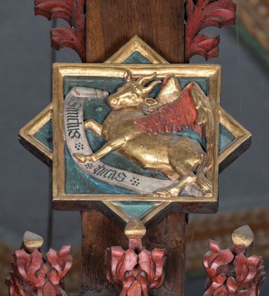 Zdjęcie nr 4: Rzeźba pełnoplastyczna, z tyłu opracowana. Ciało Chrystusa przybite do krzyża na niemal horyzontalnie, szeroko rozłożonych ramionach. Głowa w koronie cierniowej pochylona na prawe ramię. Długie włosy w puklach o spiralnie zakończonych lokach opadają na ramiona i plecy. Twarz szczupła o zindywidualizowanych rysach, pełna spokoju i smutku, okolona krótkim zarostem, z wydatnymi kośćmi policzkowymi, z kroplami krwi na czole. Duże, okrągłe oczy przesłonięte powiekami, podkreślone uniesionymi łukami brwiowymi. Usta drobne, zaciśnięte, nos prosty, wąski. Ręce chude długie, silnie wyprężone, dłonie rozchylone, przebite gwoździami. Klatka piersiowa o starannie opracowanej anatomii, z silnym wcięciem w talii oraz raną w prawym boku. Perizonium w kształcie chusty, sięgające do połowy uda, miękko udrapowane z przodu, spływające po obu bokach kaskadowymi fałdami. Nogi wyprostowane, umięśnione, ułożone obok siebie, stopy stosunkowo duże, skrzyżowane, założone prawa na lewą, przebite jednym gwoździem. Z ran na boku, dłoniach i stopach spływa obficie krew. Karnacja Chrystusa jasnoróżowa, włosy brązowe z domalowanym warkoczem z tyłu, korona zielona, perizonium złocone, z niebieską podszewką. Krzyż łaciński, dębowy, w naturalnym kolorze drewna, wykonany w 1890, w formie Drzewa Życia, z rozłożystymi liśćmi uschniętego akantu w kolorze czerwonym i złotym żyłkowaniem na skrzyżowaniu ramion. W zakończeniu ramion lilijki czerwono-zielone ze złoconymi obwódkami oraz nakładane plakiety w układzie koncentrycznym tworzące ośmioramienne gwiazdy z płaskorzeźbionymi symbolami Ewangelistów. Tła plakiet malowane na niebiesko, symbole i obramienia złocone. Plakiety współczesne z rzeźbą Chrystusa, gotyckie - 1433 r.; u góry krzyża - symbol św. Jana - orzeł, u dołu - symbol św. Łukasza - wół, na prawym ramieniu symbol św. Mateusza - anioł, na lewym ramieniu - symbol św. Marka - lew. Ponad głową Chrystusa titulus z napisem: INRI. Plakieta kwadratowa nałożona na romb w układzie koncentrycznym, w profilowanym obramieniu, z płaskorzeźbionym przedstawieniem orła; u dołu banderola z napisem minuskułą gotycką: sanctus joannes. Plakieta kwadratowa nałożona na romb w układzie koncentrycznym, w obramieniu profilowanym, z fazą wklęsłą - z płaskorzeźbionym przedstawieniem anioła w pozycji siedzącej z podkurczonymi nogami w ujęciu frontalnym, w rękach podtrzymującego faliście wygiętą banderolę o zrolowanych zakończeniach, z czarnym napisem minuskułą gotycką: sanctus matheus; banderola biała z czarnymi rozetkami. Twarz anioła okrągła, o rysach dziecka, różowa, okolona puklami rudych włosów. Anioł ubrany w fałdzistą szatę, złoconą z trójkątnym dekoltem, udrapowaną na kolanach w głębokie, łyżkowate fałdy. U ramion szeroko rozpostarte skrzydła o schematycznych rombowych podziałach, zagięte na brzegach, zakończone długimi piórami, lotkami - złocone. Tło lekko pogłębione, niebieskie; obramienia kwadratów złocone, boki i tylne ściany malowane na brązowo. Plakieta kwadratowa nałożona na romb w układzie koncentrycznym, w obramieniu profilowanym, z fazą wklęsłą, z płaskorzeźbionym przedstawieniem uskrzydlonego wołu, w pełnej postaci, w ujęciu bocznym, o masywnej budowie ciała, w dynamicznym ruchu, wspartego na rozstawionych nogach, zakończonych ciężkimi kopytami, z puszystym ogonem. Głowa zwrócona na wprost, oczy duże, nozdrza rozdęte, uszy odstające na boki, pośrodku ozdobnie osadzone esowato wygięte rogi. Skrzydła wyrastające z barków, uniesione w górę, rozpostarte, upierzenie schematyczne zaznaczone w kształcie rombów, z długimi lotkami w zakończeniach wygiętych skrzydeł. U dołu, pomiędzy nogami zwierzęcia - półkoliście wygięta biała banderola, z czarnym napisem minuskułą gotycką: sanctus lucas i czarnymi rozetkami. Tło nieco pogłębione, niebieskie. Postać wołu, obramienie plakiet, złocone; ścianki tylne malowane na brązowo. Plakieta w kształcie rombu nałożonego na kwadrat w układzie koncentrycznym, w obramieniu profilowanym, z fazą wklęsłą - z płaskorzeźbionym przedstawieniem uskrzydlonego lwa. W pełnej postaci, ujęciu bocznym, muskularnych kształtach, mocnych nogach z potężnymi pazurami. Głowa zwrócona na wprost, ujęta bujną, silnie skręconą grzywą. Skrzydła wyrastające z barków uniesione w górę, szeroko rozpostarte, zakończone długimi lotkami; upierzenie schematycznie zaznaczone w formie regularnych rombów. Ogon cienki, długi, z puszystą kitką, przechodzący pomiędzy nogami, esowato wygięty nad grzbietem. U dołu biała banderola, podtrzymywana pazurami przednich łap, z napisem czarnym, gotycką minuskułą: sanctus marcus i czarnymi rozetkami. Tło nieco pogłębione, niebieskie; postać lwa i obramienia złocone, tylne ścianki malowane na brązowo.