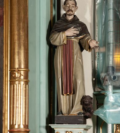 Zdjęcie nr 6: Rzeźba pełnoplastyczna, ustawiona na podstawie na planie prostokąta. Święty Dominik ukazany frontalnie, w kontrapoście, z pochyloną głową, prawą rękę kładzie na piersi, w lewej wyciągniętej do przodu trzyma różaniec. Twarz o wyraźnie zaznaczonych zmarszczkach, dużych uszach, zapadniętych policzkach, zmarszczonym czole, o dużych oczach skierowanych w dół, wydatnym nosie i wąskich ustach z gęstymi wąsami. Włosy ma krótkie, brązowe, z tonsurą na głowie. Święty ubrany jest w biały habit ze złoconą lamówką, przepasany w talii, z różowym szkaplerzem, na ramiona ma narzuconą brązową pelerynkę z kapturem i złoconą lamówką. Polichromia ciała naturalistyczna, o bladej karnacji. Przy lewej nodze świętego postać brązowego, leżącego psa. 



