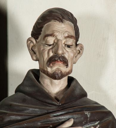 Zdjęcie nr 1: Rzeźba pełnoplastyczna, ustawiona na podstawie na planie prostokąta. Święty Dominik ukazany frontalnie, w kontrapoście, z pochyloną głową, prawą rękę kładzie na piersi, w lewej wyciągniętej do przodu trzyma różaniec. Twarz o wyraźnie zaznaczonych zmarszczkach, dużych uszach, zapadniętych policzkach, zmarszczonym czole, o dużych oczach skierowanych w dół, wydatnym nosie i wąskich ustach z gęstymi wąsami. Włosy ma krótkie, brązowe, z tonsurą na głowie. Święty ubrany jest w biały habit ze złoconą lamówką, przepasany w talii, z różowym szkaplerzem, na ramiona ma narzuconą brązową pelerynkę z kapturem i złoconą lamówką. Polichromia ciała naturalistyczna, o bladej karnacji. Przy lewej nodze świętego postać brązowego, leżącego psa. 



