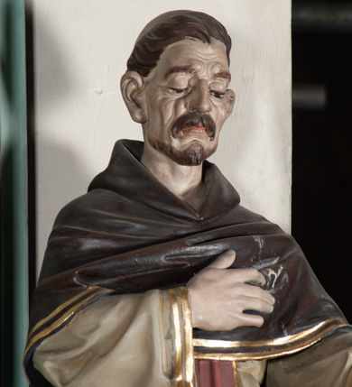 Zdjęcie nr 4: Rzeźba pełnoplastyczna, ustawiona na podstawie na planie prostokąta. Święty Dominik ukazany frontalnie, w kontrapoście, z pochyloną głową, prawą rękę kładzie na piersi, w lewej wyciągniętej do przodu trzyma różaniec. Twarz o wyraźnie zaznaczonych zmarszczkach, dużych uszach, zapadniętych policzkach, zmarszczonym czole, o dużych oczach skierowanych w dół, wydatnym nosie i wąskich ustach z gęstymi wąsami. Włosy ma krótkie, brązowe, z tonsurą na głowie. Święty ubrany jest w biały habit ze złoconą lamówką, przepasany w talii, z różowym szkaplerzem, na ramiona ma narzuconą brązową pelerynkę z kapturem i złoconą lamówką. Polichromia ciała naturalistyczna, o bladej karnacji. Przy lewej nodze świętego postać brązowego, leżącego psa. 



