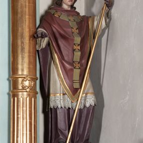 Zdjęcie nr 1: Rzeźba pełnoplastyczna, ustawiona na podstawie na planie prostokąta. Święty Stanisław ukazany frontalnie, w kontrapoście, w lewej uniesionej z przodu ręce trzyma pastorał, prawą wyciąga przed siebie. Twarz ma szczupłą, o podkreślonym zmarszczonym czole i brwiach, wystających kościach policzkowych i brodzie; o migdałowatych, półprzymkniętych oczach, krótkim nosie i wąskich ustach. Włosy ma brązowe, kręcone, opadające na ramiona. Ubrany jest w fioletową albę, białą rokietę z motywem koronki i złoconej lamówki przy brzegach, bordową kapę obwiedzioną złoconą i zieloną lamówką, na piersi ma jasnozielony paliusz ze złoconymi krzyżami na czarnym tle, na głowie ma białą infułę, ze złoconą lamówka, na dłoniach fioletowe rękawiczki, a na stopach czarne buty. Polichromia ciała naturalistyczna, o bladej karnacji. 
