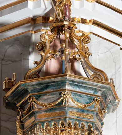 Zdjęcie nr 6: Ambona przyścienna, wisząca ustawiona przy zachodnim filarze łuku tęczowego; dostępna jednobiegowymi schodami z balustradą od strony prezbiterium. Kosz na planie ośmioboku, na puklowanej podstawie spiętej u dołu złoconą obręczą z kwiatem. U dołu wydzielony silnie występującym ćwierćwałkiem z nałożoną złoconą dekoracją w formie liści akantu;  u góry zamknięty profilowanym gzymsem.  Ścianki artykułowane łodygą roślinną o pionowym przebiegu. W polach prostokątne płyciny mieszczące płaskorzeźbione portrety czterech ewangelistów wraz z ich atrybutami umieszczone na tle symetrycznego  ornamentu roślinnego. Balustrada schodów pełna; pola flankowane analogiczną jak na koszu łodygą, pośrodku pól dwie wielolistne rozety. Zaplecek flankowany jońskimi kolumnami o złoconych, kanelowanych trzonach wspierających baldachim oraz uszami utworzonymi z rocaille'ów, kratki regencyjnej oraz liści laurowych. W polu prostokątna płycina zamknięta łukiem wklęsło-wypukłym z uskokiem mieszcząca obraz Dobrego Pasterza malowany olejem na desce. Baldachim ośmioboczny, profilowany z podwieszonym u dołu lambrekinem. W partii fryzu girlandy podwieszone na kokardach z wstęgi regencyjnej. W zwieńczeniu ażurowa  struktura utworzona z wolut wspierająca postument z pełnoplastyczną, polichromowaną figurą św. Pawła.
Polichromia jasnobeżowa, miejscami marmoryzowana w odcieniach morskiego błękitu; ornamentyka oraz detale architektoniczne złocone i srebrzone.