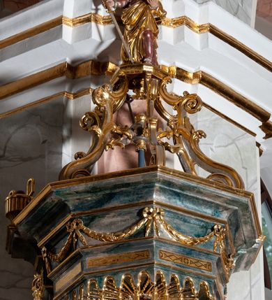 Zdjęcie nr 5: Ambona przyścienna, wisząca ustawiona przy zachodnim filarze łuku tęczowego; dostępna jednobiegowymi schodami z balustradą od strony prezbiterium. Kosz na planie ośmioboku, na puklowanej podstawie spiętej u dołu złoconą obręczą z kwiatem. U dołu wydzielony silnie występującym ćwierćwałkiem z nałożoną złoconą dekoracją w formie liści akantu;  u góry zamknięty profilowanym gzymsem.  Ścianki artykułowane łodygą roślinną o pionowym przebiegu. W polach prostokątne płyciny mieszczące płaskorzeźbione portrety czterech ewangelistów wraz z ich atrybutami umieszczone na tle symetrycznego  ornamentu roślinnego. Balustrada schodów pełna; pola flankowane analogiczną jak na koszu łodygą, pośrodku pól dwie wielolistne rozety. Zaplecek flankowany jońskimi kolumnami o złoconych, kanelowanych trzonach wspierających baldachim oraz uszami utworzonymi z rocaille'ów, kratki regencyjnej oraz liści laurowych. W polu prostokątna płycina zamknięta łukiem wklęsło-wypukłym z uskokiem mieszcząca obraz Dobrego Pasterza malowany olejem na desce. Baldachim ośmioboczny, profilowany z podwieszonym u dołu lambrekinem. W partii fryzu girlandy podwieszone na kokardach z wstęgi regencyjnej. W zwieńczeniu ażurowa  struktura utworzona z wolut wspierająca postument z pełnoplastyczną, polichromowaną figurą św. Pawła.
Polichromia jasnobeżowa, miejscami marmoryzowana w odcieniach morskiego błękitu; ornamentyka oraz detale architektoniczne złocone i srebrzone.